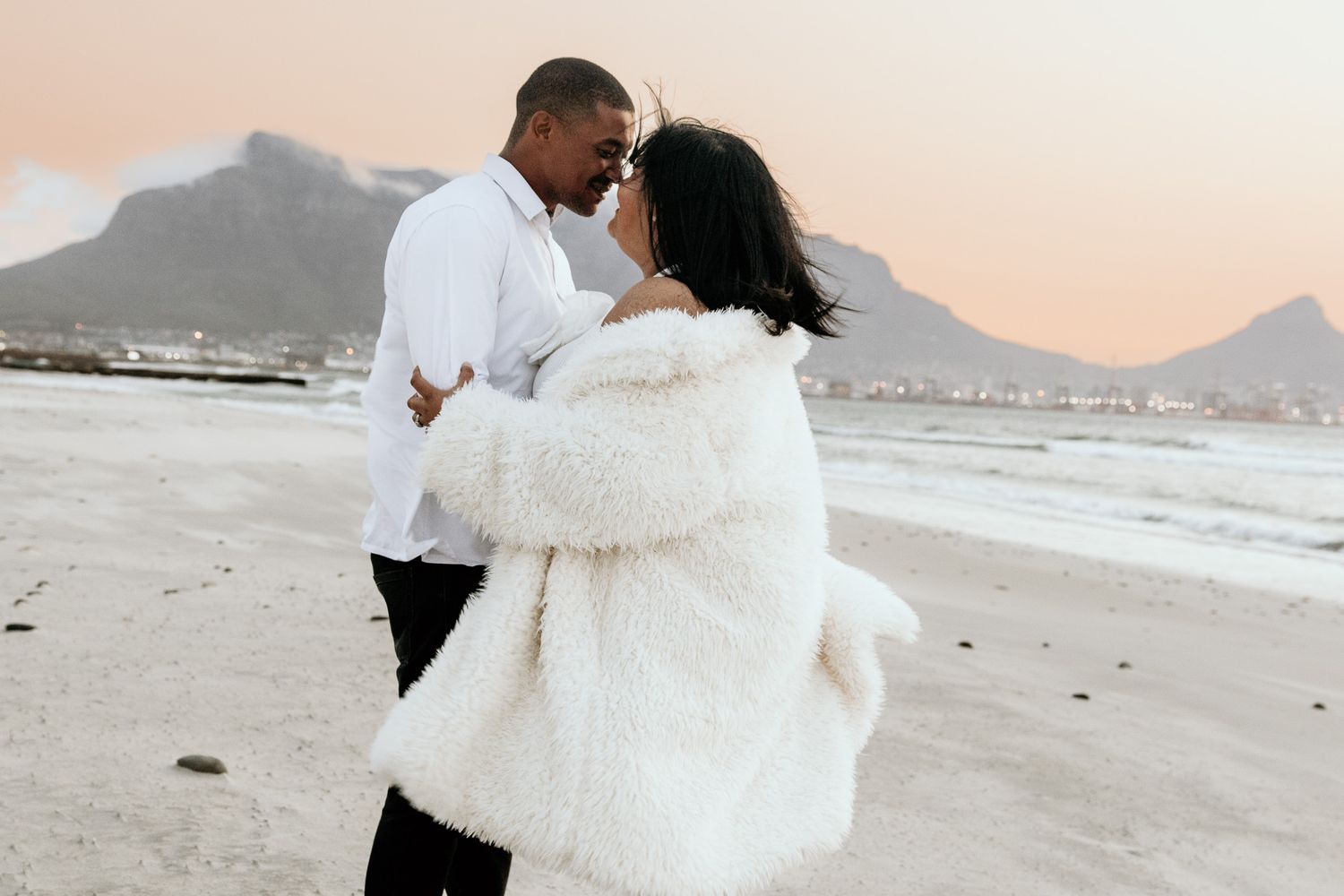 Couple embracing on beach at sunset with mountain backdrop wearing white outfits.