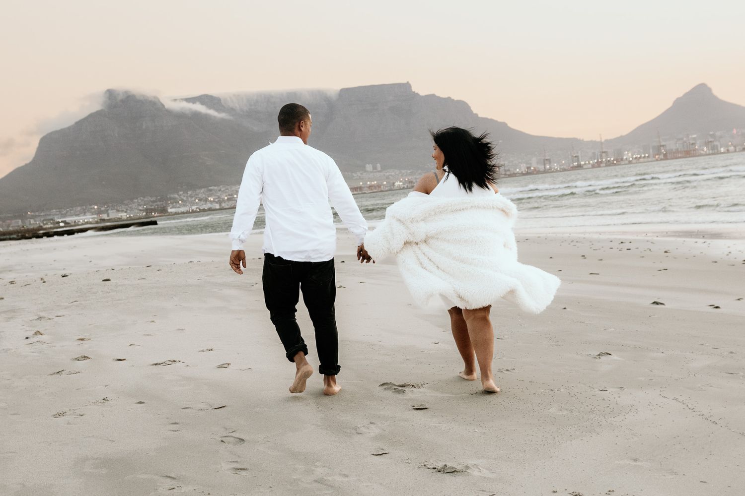 Couple in white outfits running hand-in-hand on a sandy beach with Table Mountain backdrop.