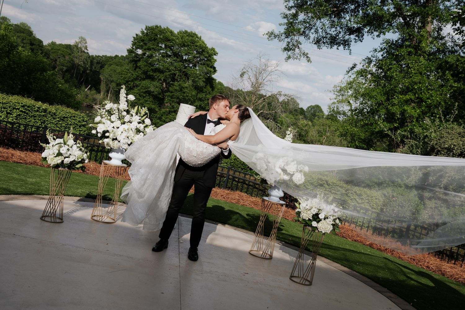 Groom sweeps bride off her feet as her long veil flows dramatically at outdoor garden wedding.