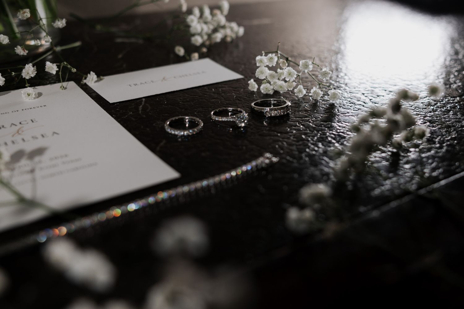 Wedding flat lay with invitation, diamond rings, bracelet, and delicate baby's breath flowers on dark surface.