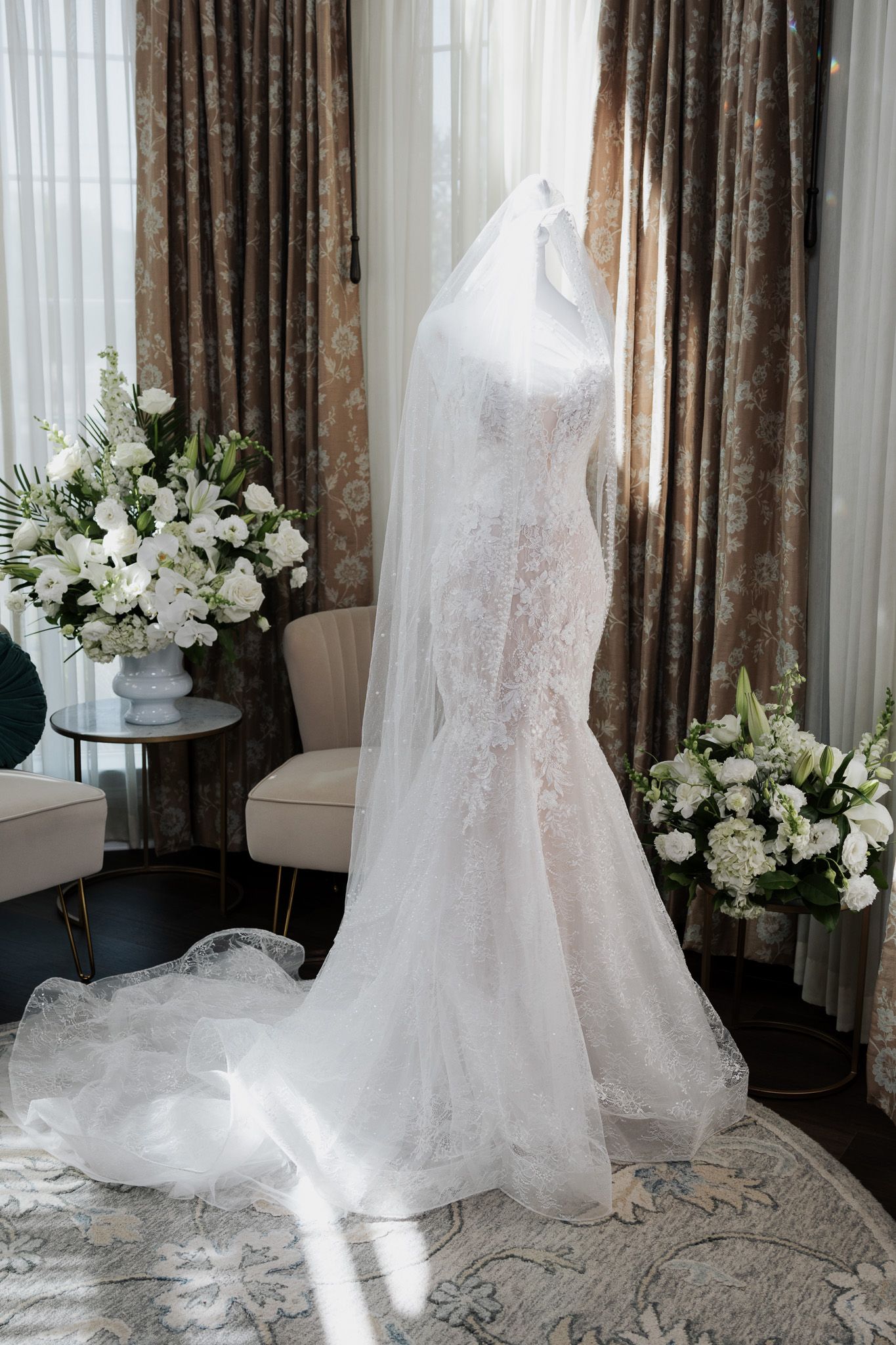 Elegant lace mermaid wedding dress with long veil displayed on mannequin by sunlit window.
