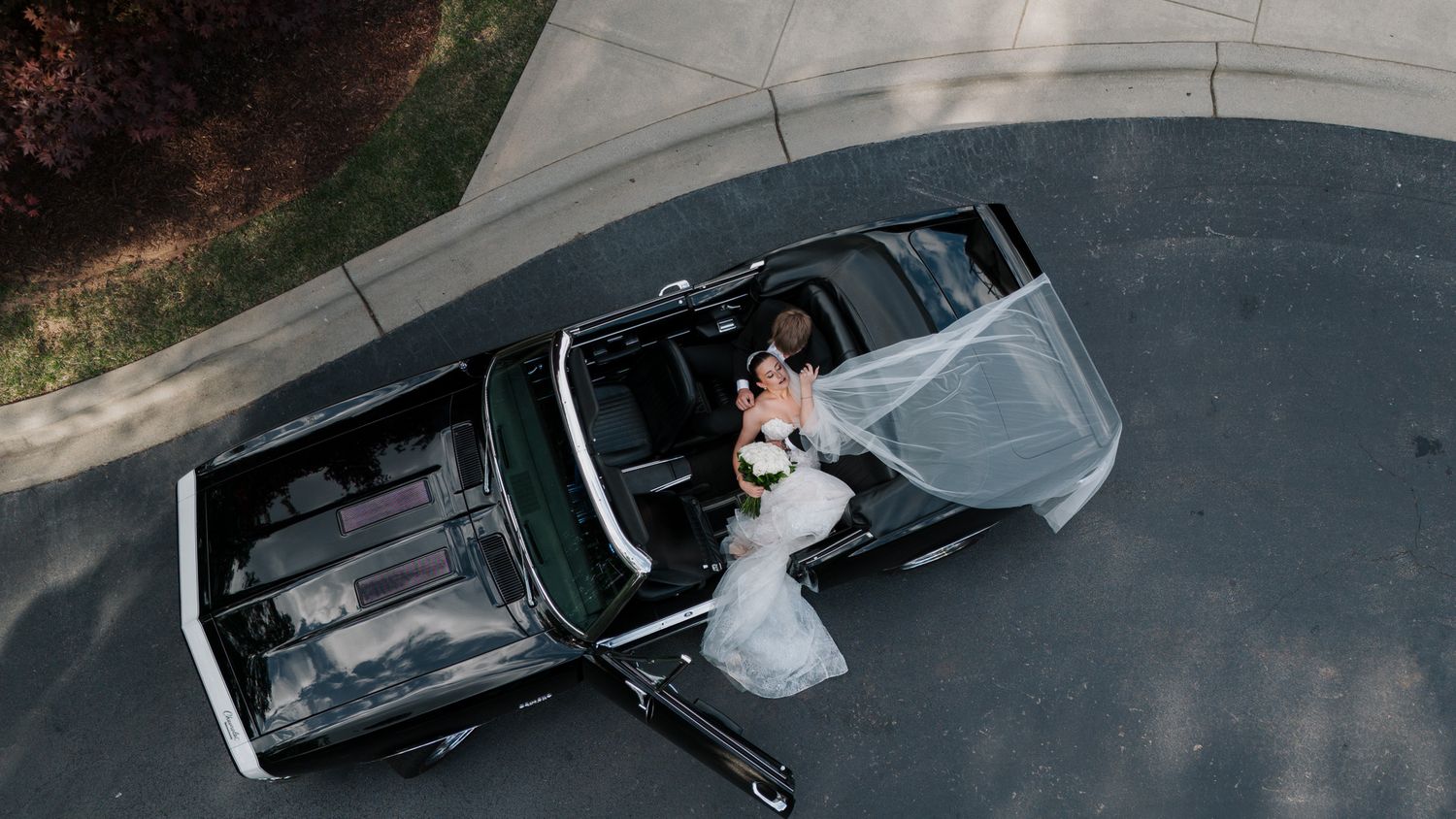 Drone overhead shot of bride and groom kissing beside a vintage black convertible car.