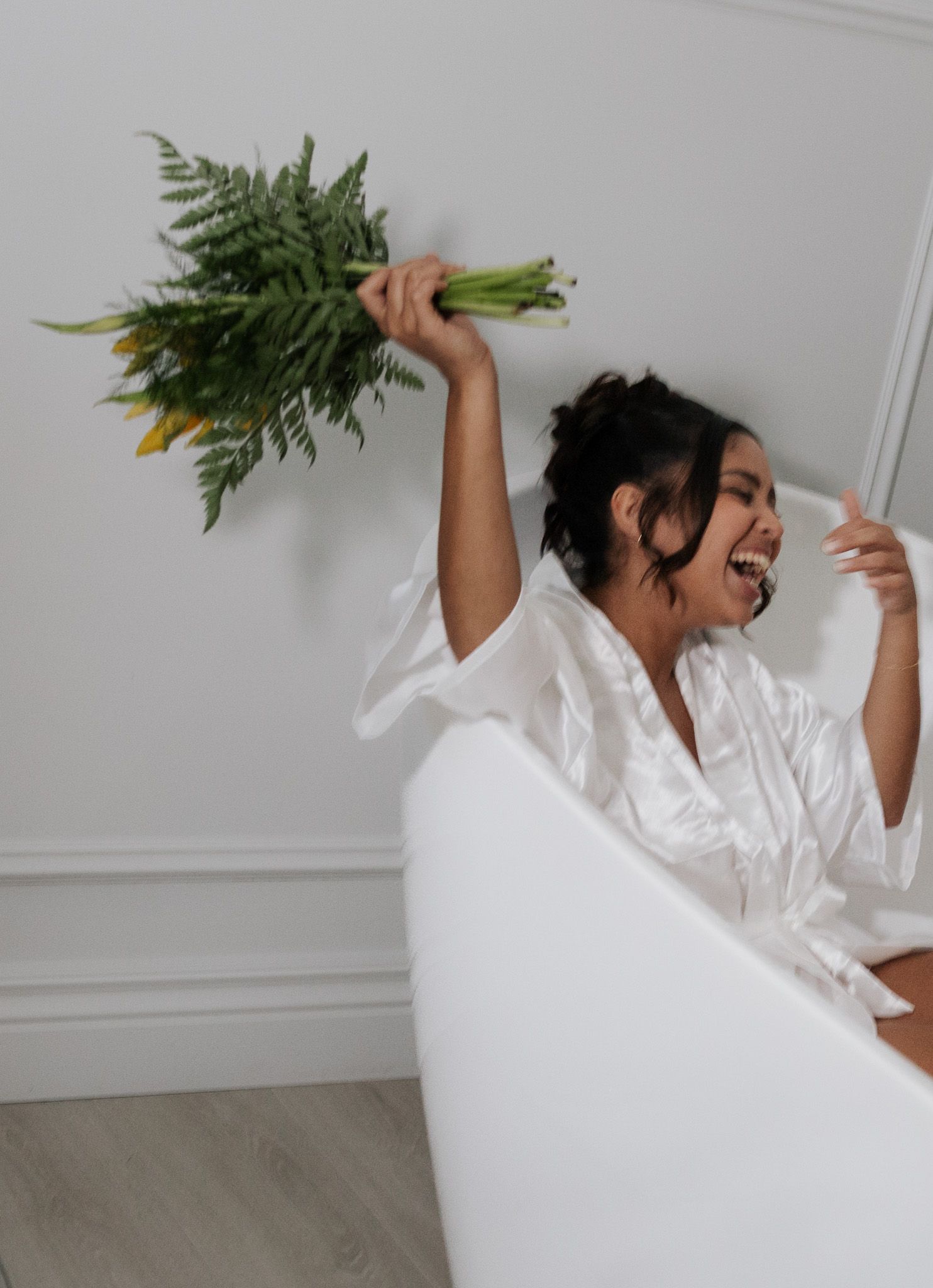 Joyful bridesmaid in white robe holds green bouquet aloft while laughing on a white sofa.
