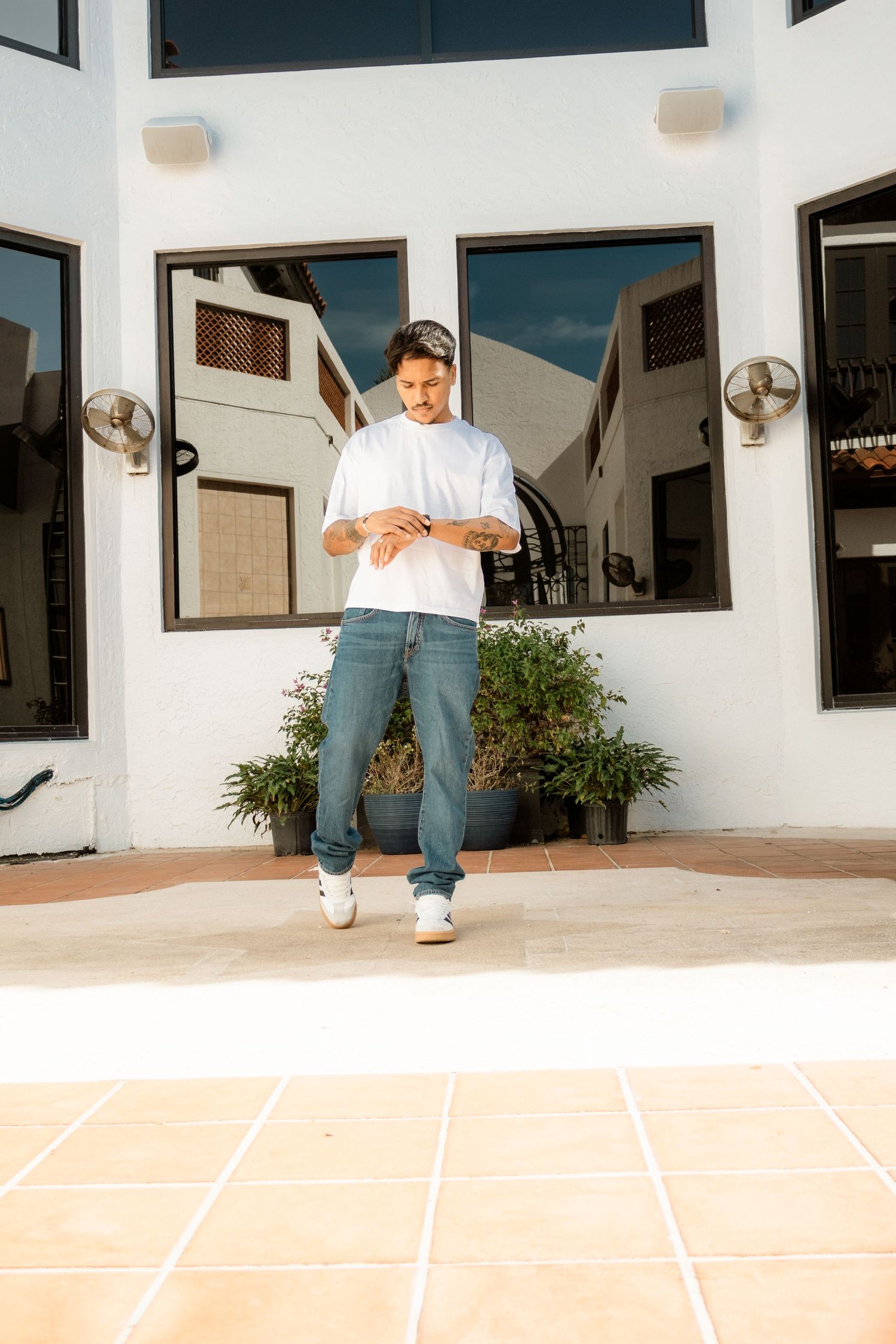 Man in white t-shirt and jeans checking his watch outside a modern white building.