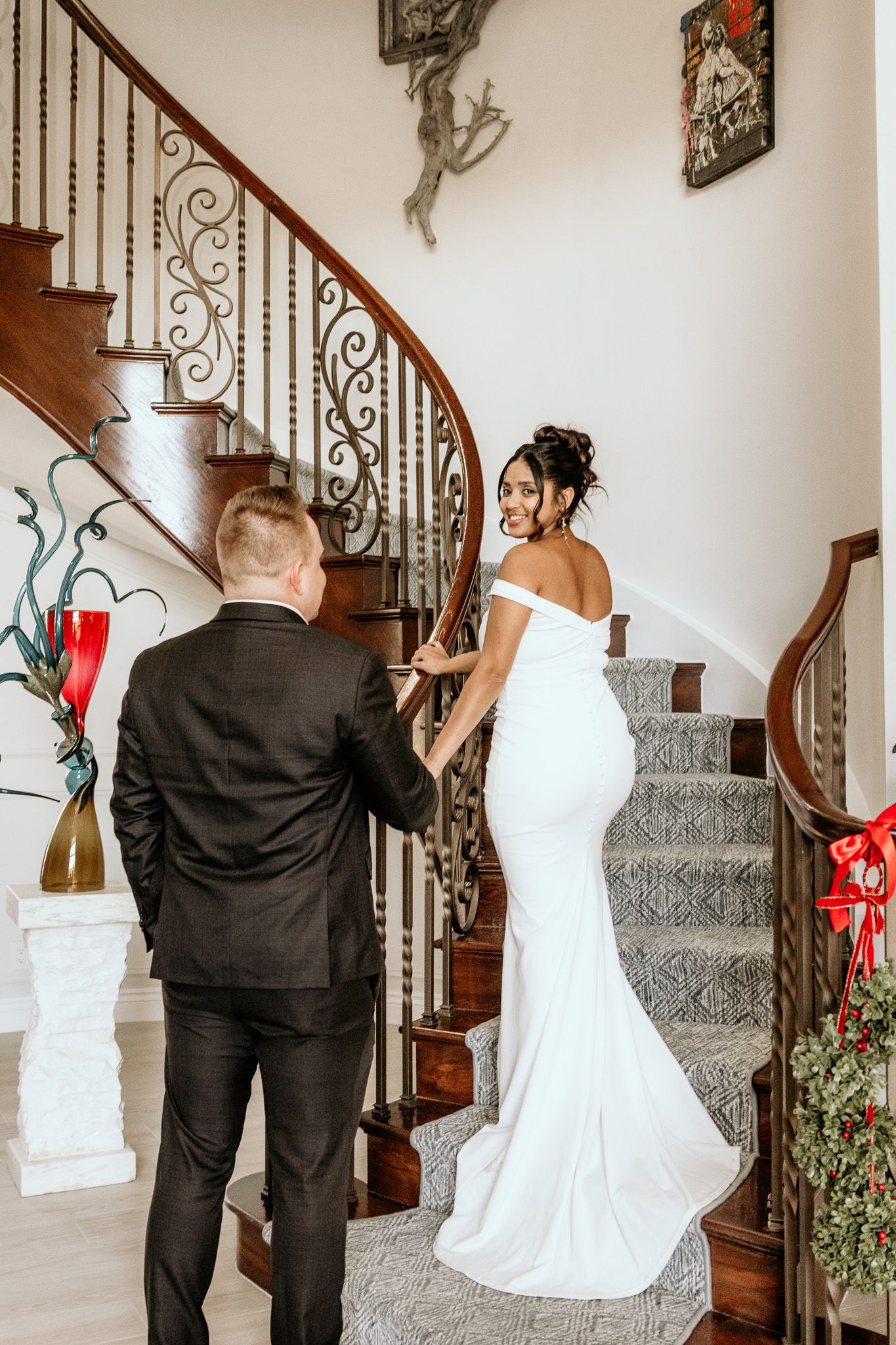 Bride in fitted white gown smiling on curved staircase with groom in black suit.