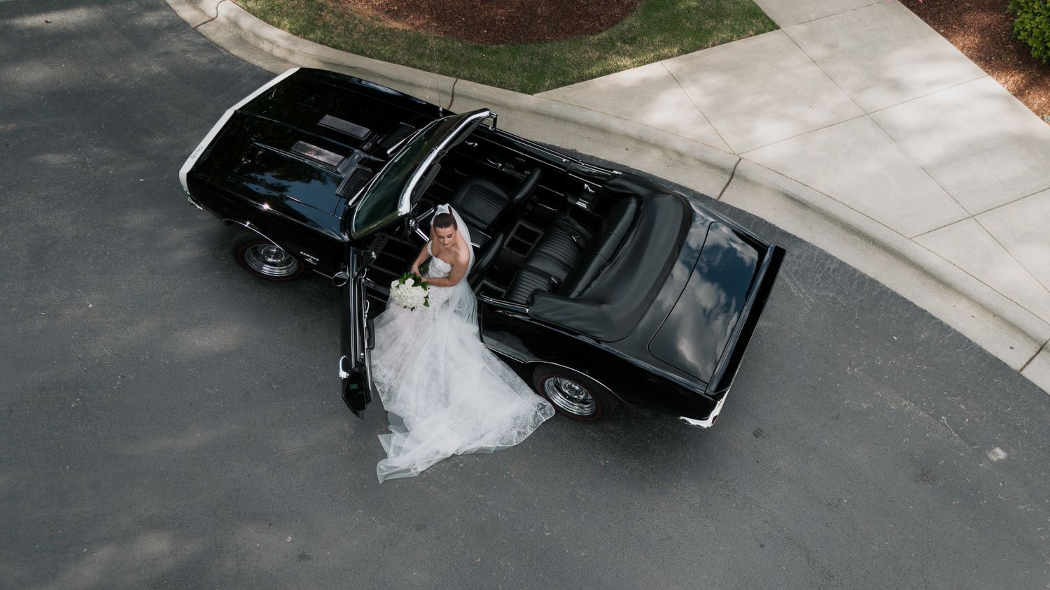 Aerial view of bride in white wedding gown standing beside a classic black convertible car.