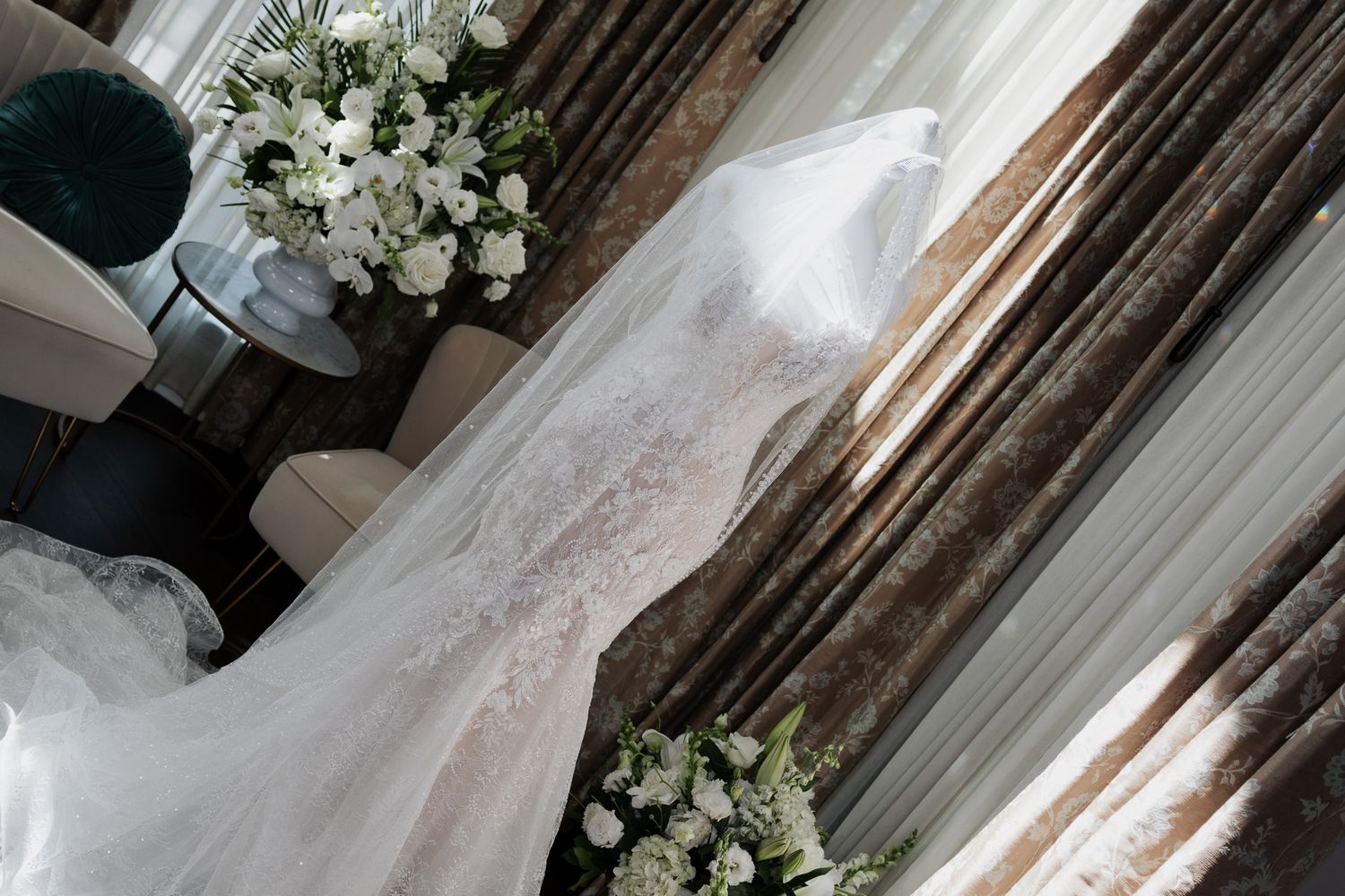Elegant lace wedding dress displayed near a window with white floral bouquets nearby.