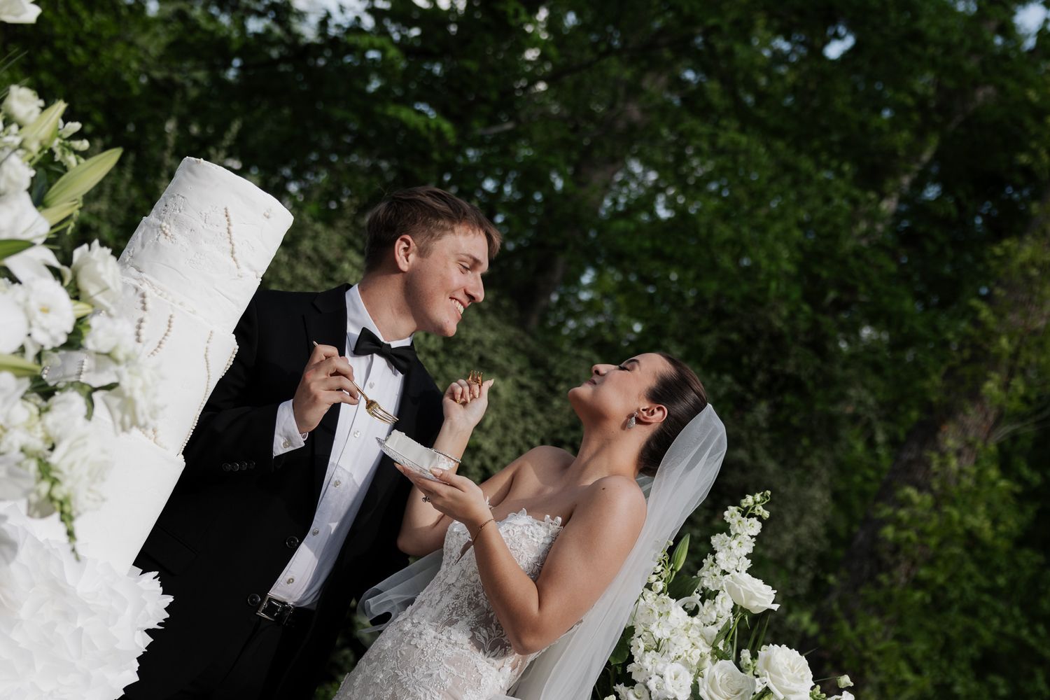 Smiling bride and groom cutting their white tiered wedding cake outdoors among beautiful white flower arrangements.