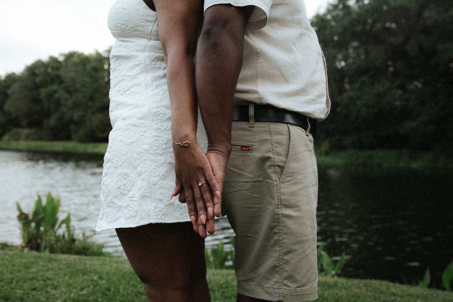 An engaged couple holds hands outdoors, showcasing a sparkling diamond engagement ring near a peaceful lake.