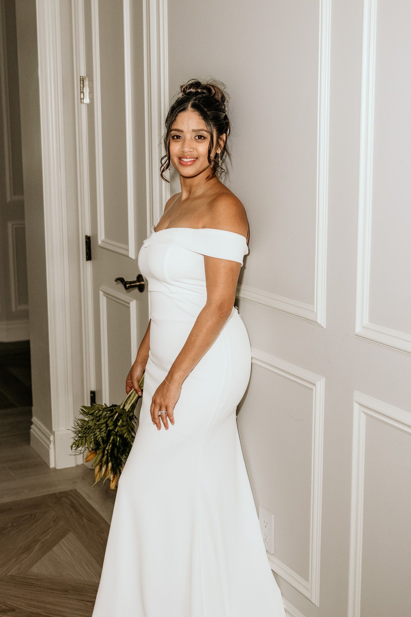 Bride in off-shoulder white mermaid gown holding yellow bouquet, smiling against white paneled wall.