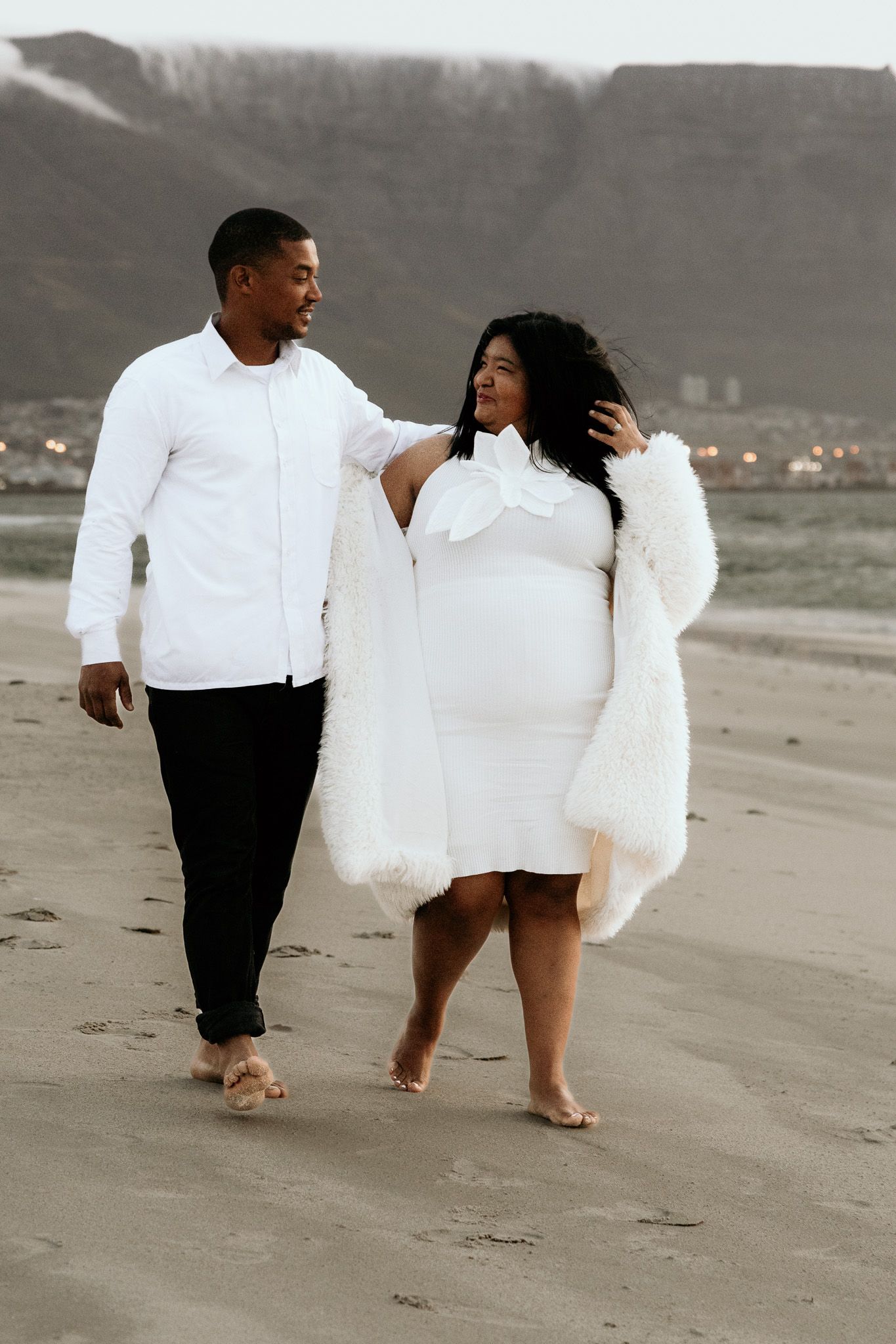 Pregnant couple in white outfits walking barefoot on a beach with mountains in the background.