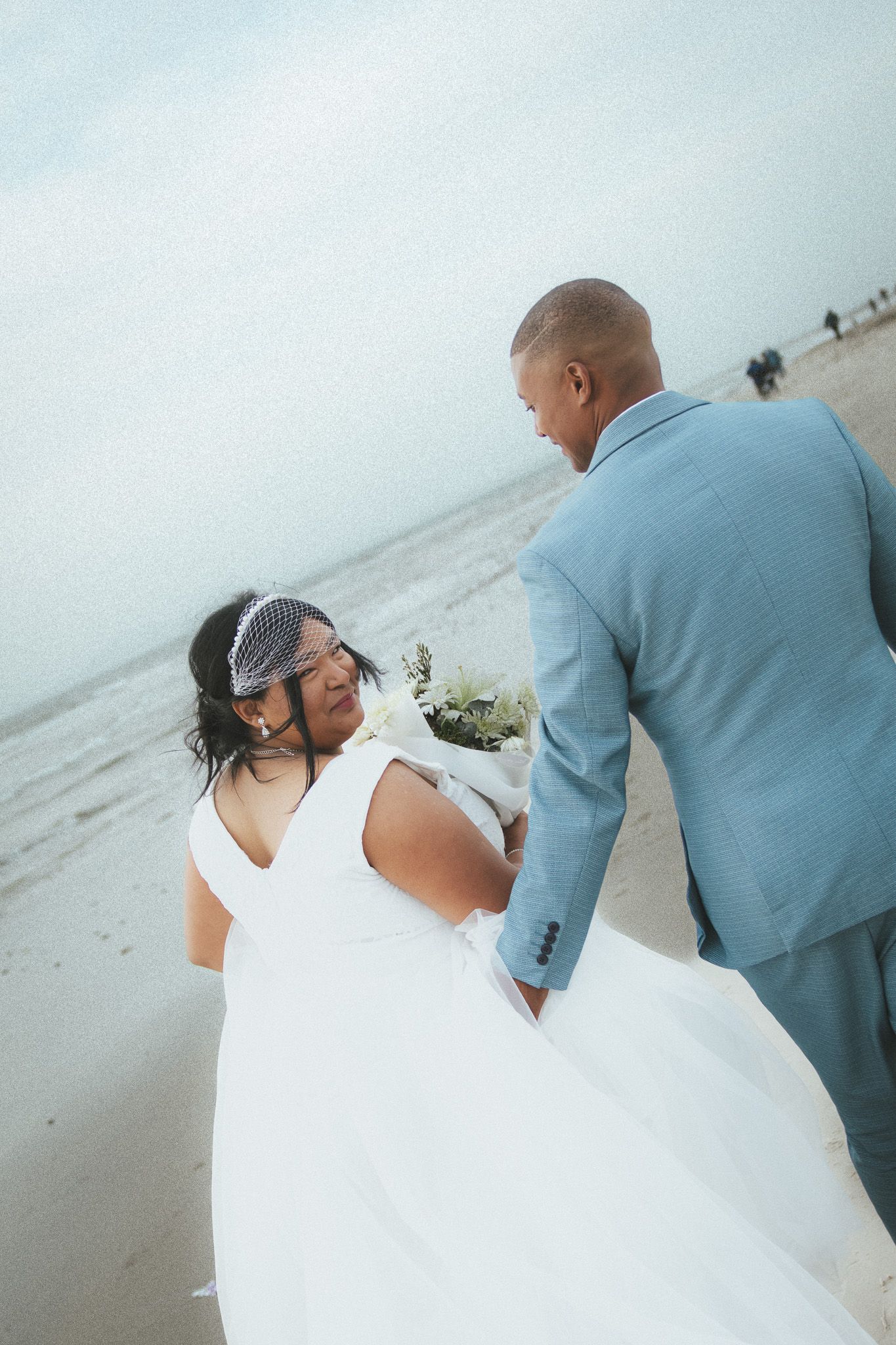 A bride and groom share a romantic moment on a beach, with the bride in a white gown and groom in a light blue suit.