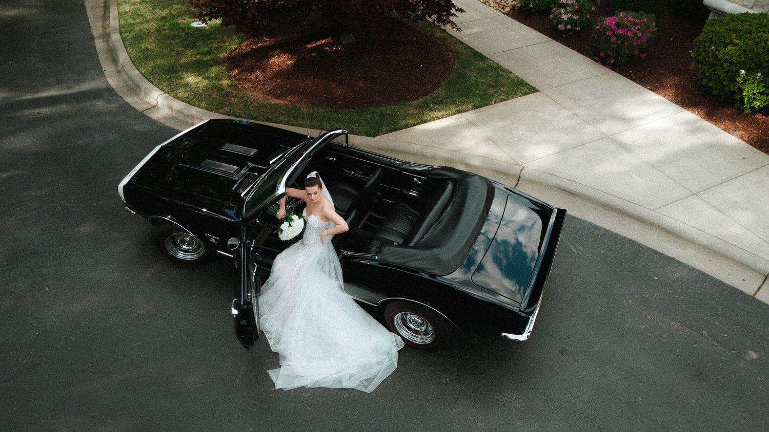 Aerial view of elegant bride in white gown posing beside a sleek black vintage convertible.