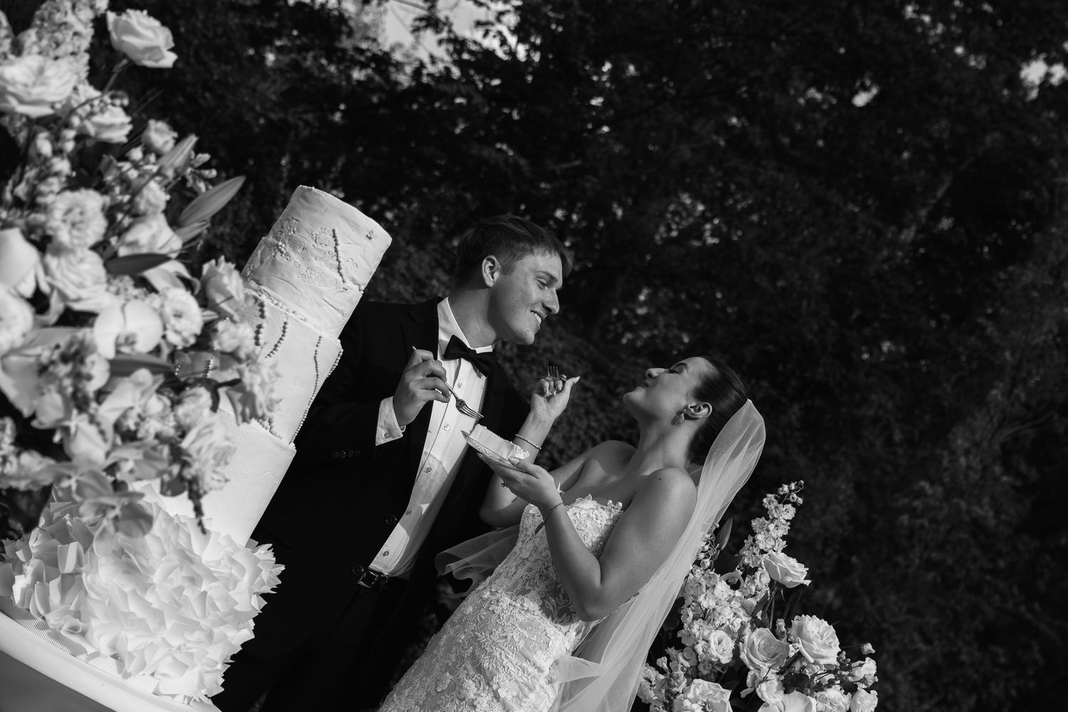 Black and white photo of bride and groom cutting their wedding cake surrounded by elegant floral decorations.