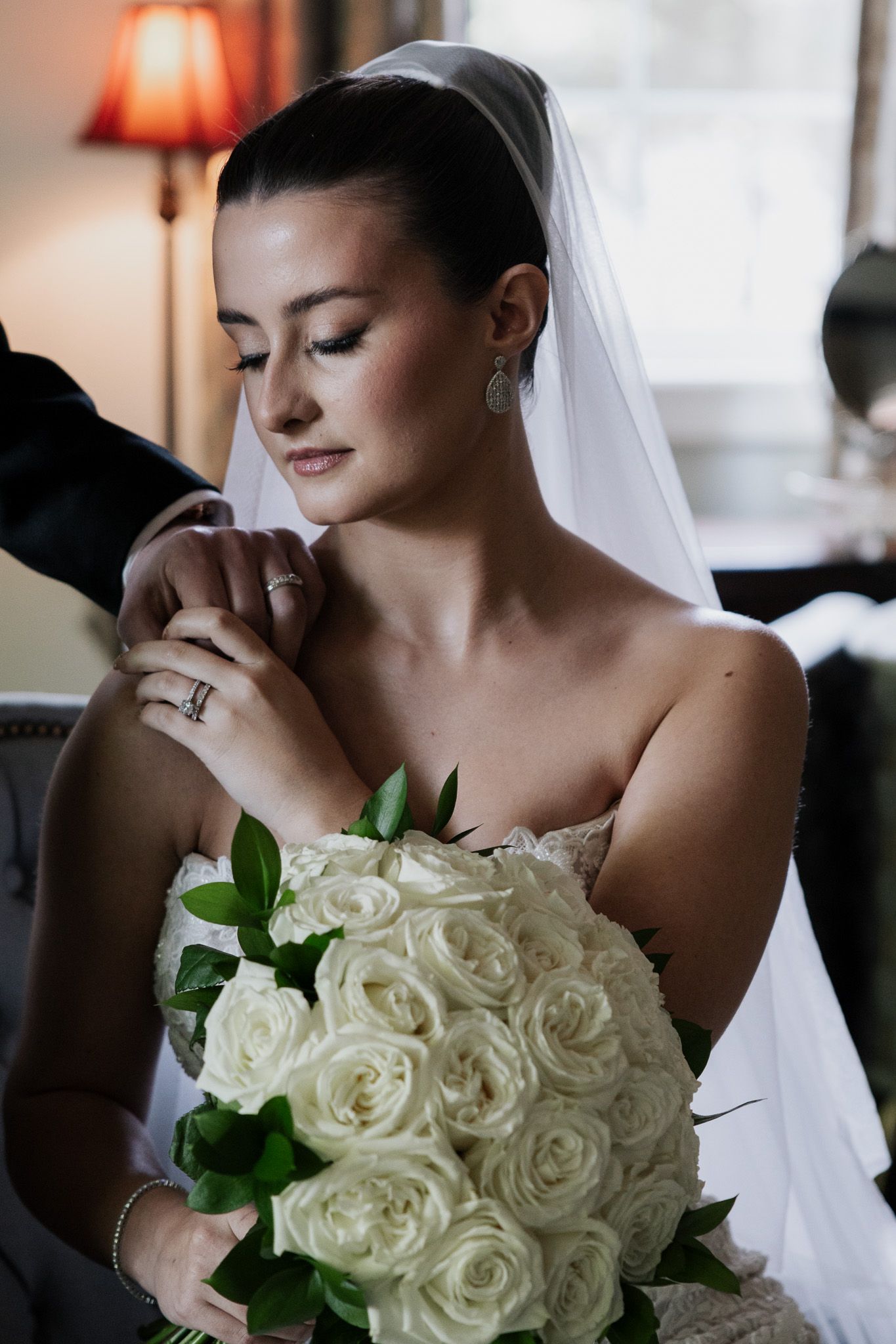 Elegant bride in strapless lace gown with veil holding white rose bouquet, eyes closed, in warm light.