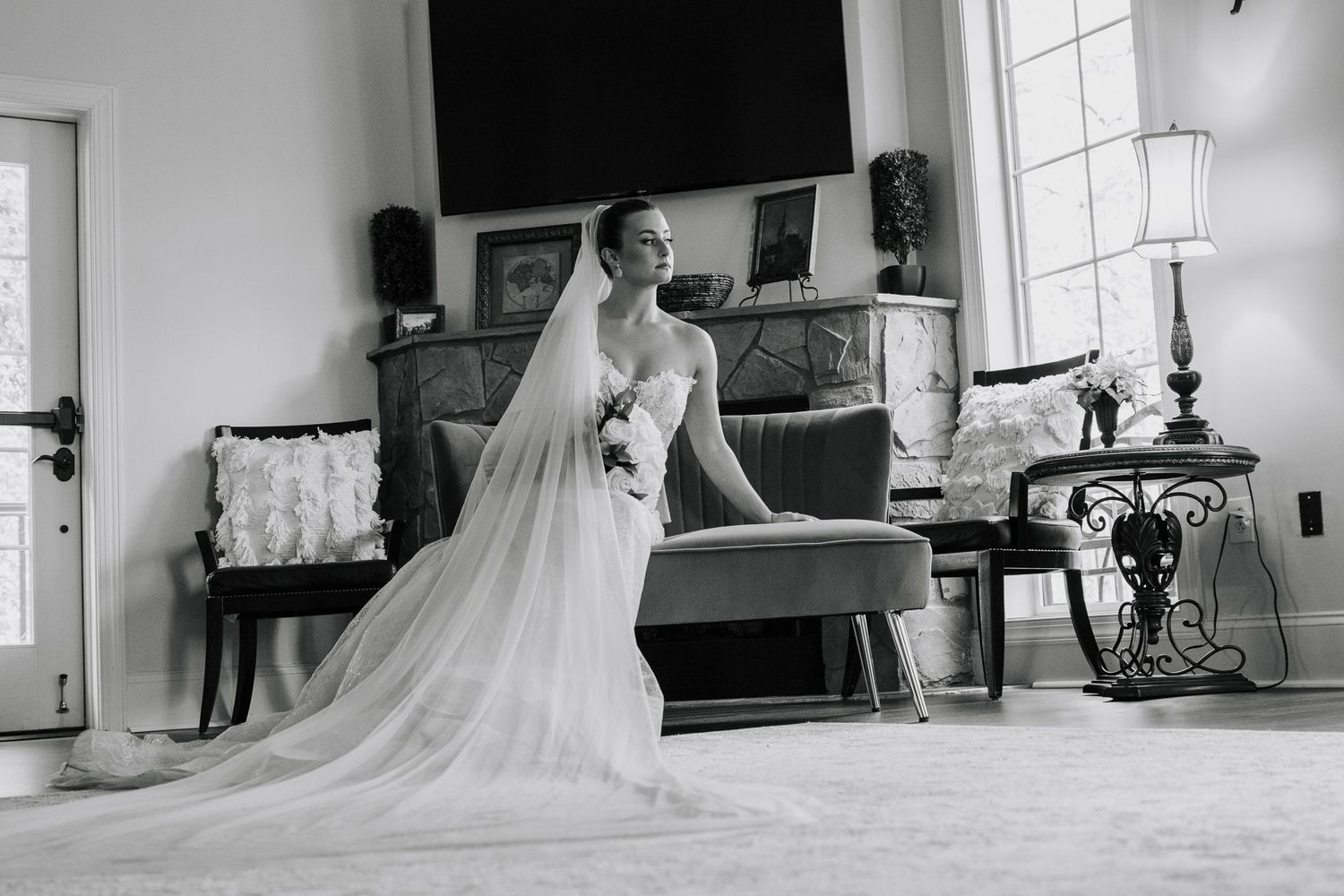 Black and white photo of a bride in a flowing wedding gown and veil standing in an elegantly furnished room.