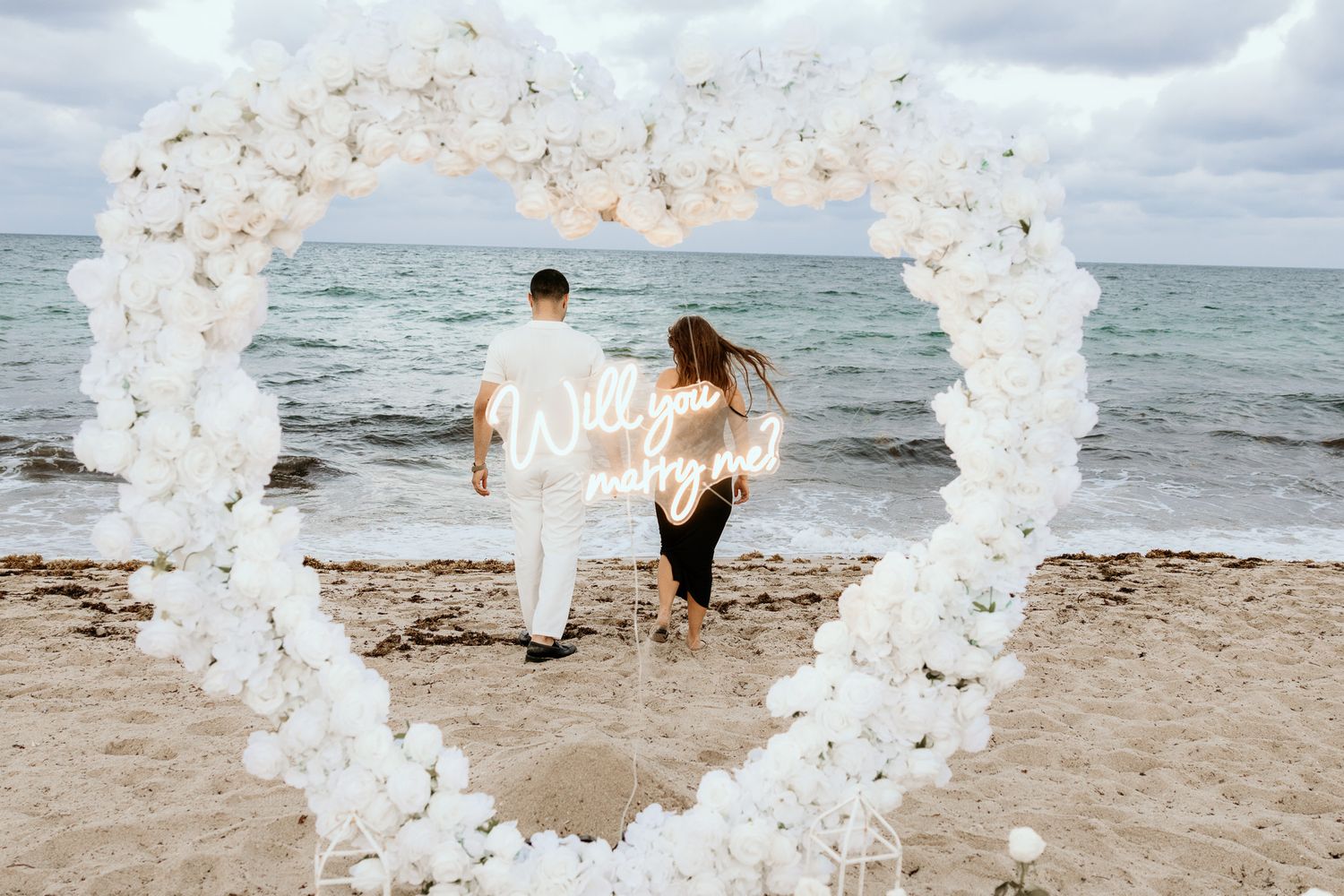 Couple stands inside a white floral heart arch on a beach with a Will You Marry Me neon sign.