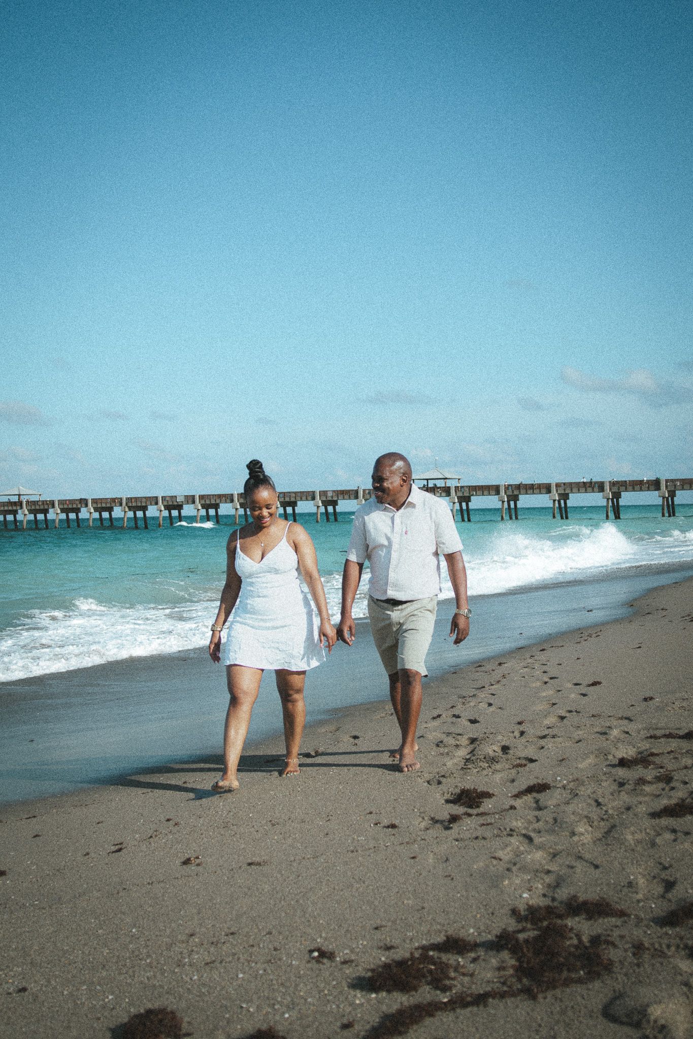 Happy couple in matching white outfits walk hand in hand along a sunny tropical beach with a pier in the background.