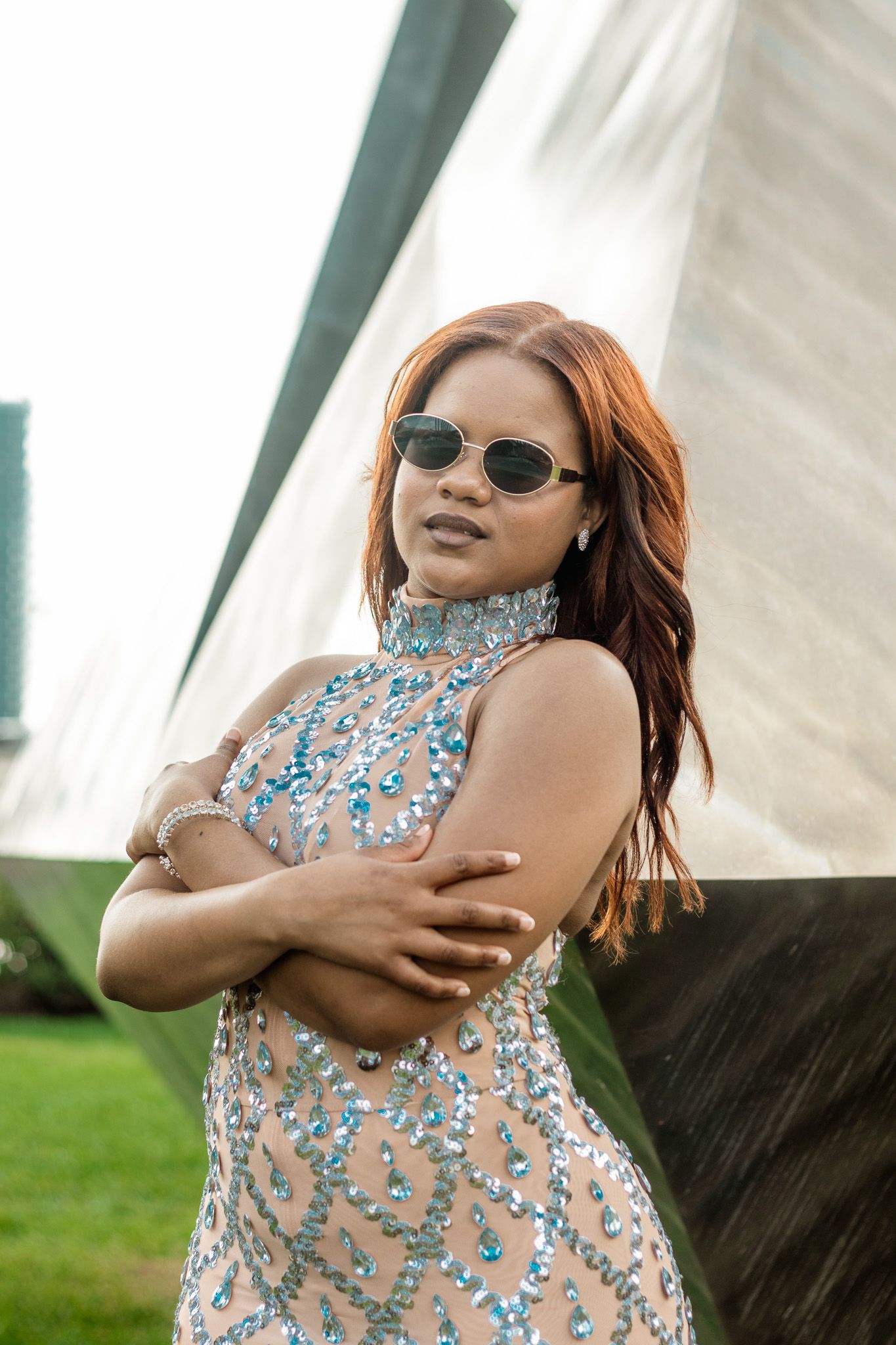 Woman in jeweled sheer dress with sunglasses poses confidently with arms crossed outdoors.