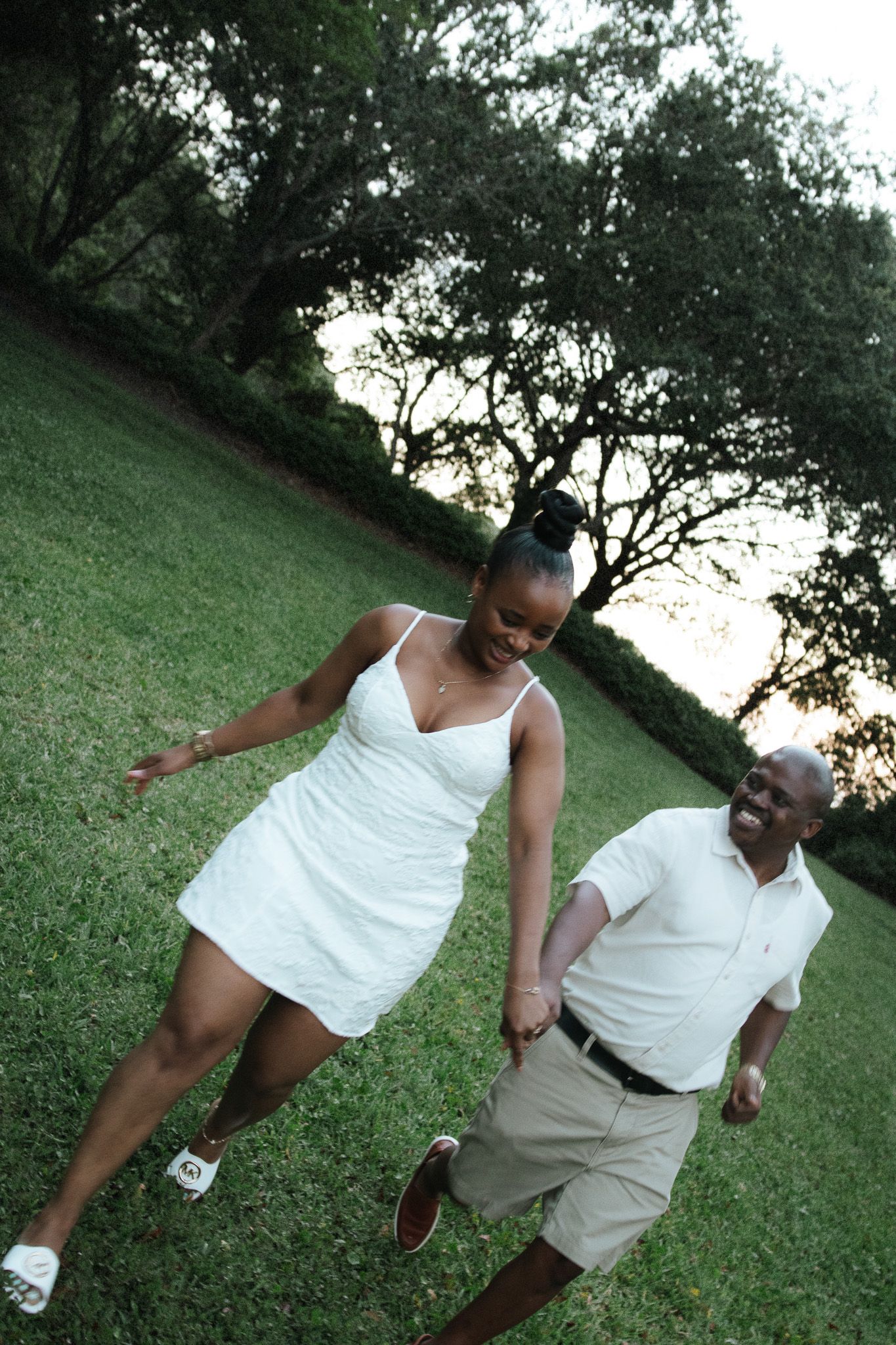 A laughing couple holds hands and walks playfully across a green lawn at golden hour in a park.