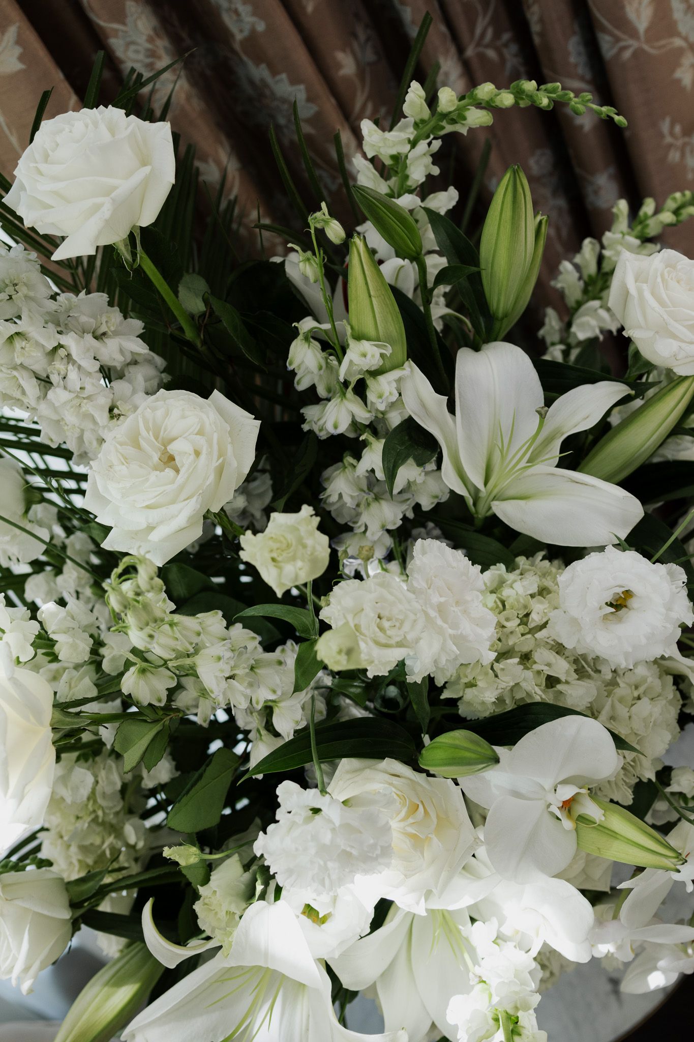 Elegant white wedding floral arrangement featuring roses, lilies, and mixed blooms against a draped backdrop.
