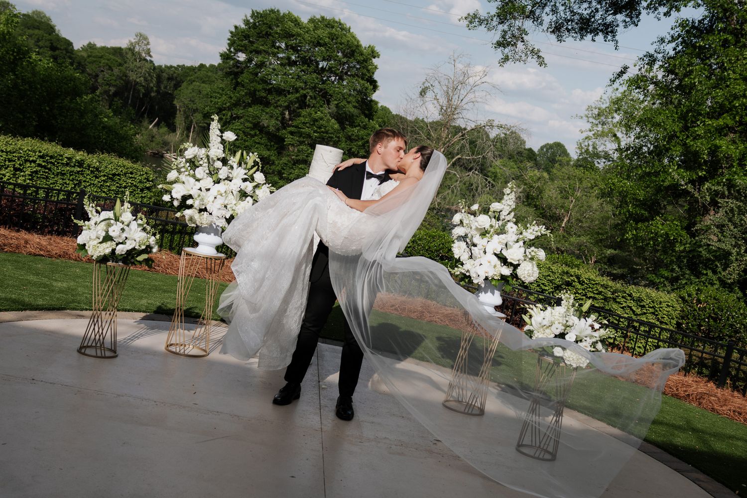 Groom lifts and dips bride in flowing white dress during outdoor wedding ceremony with white flowers.