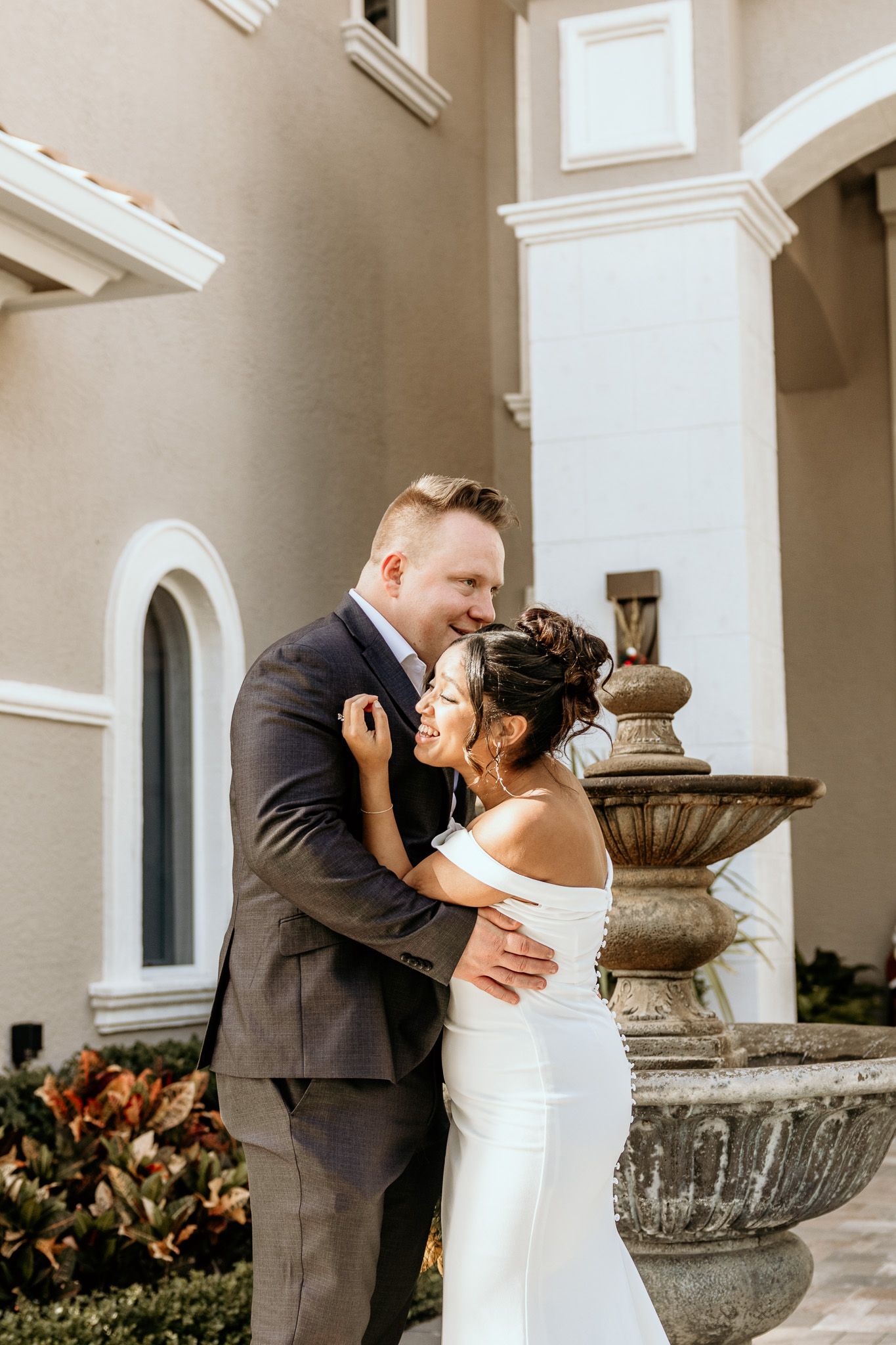 Bride and groom embracing and laughing near stone fountain outside elegant venue.