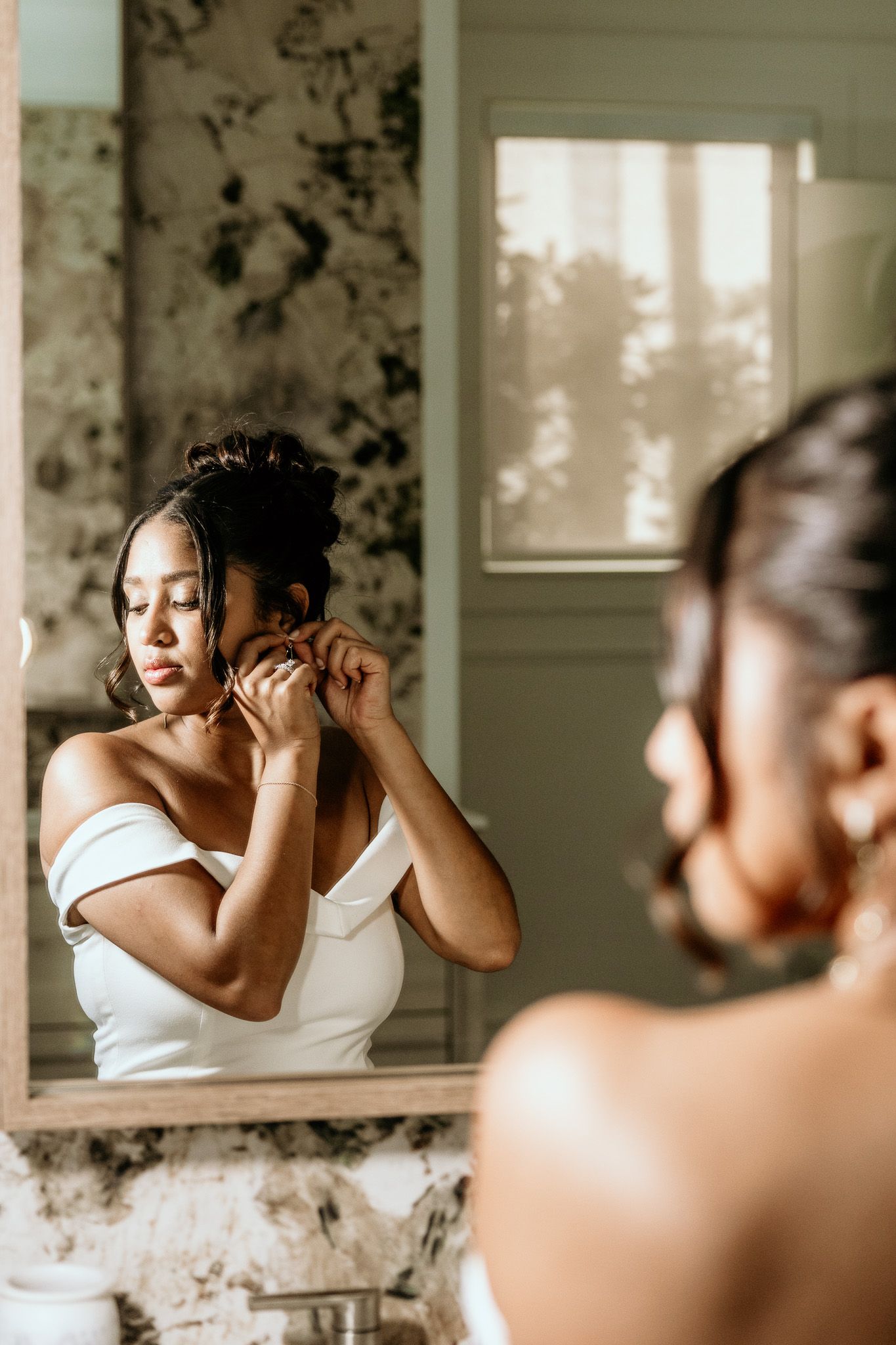 Bride putting on earrings while looking in ornate bathroom mirror wearing off-shoulder white gown.