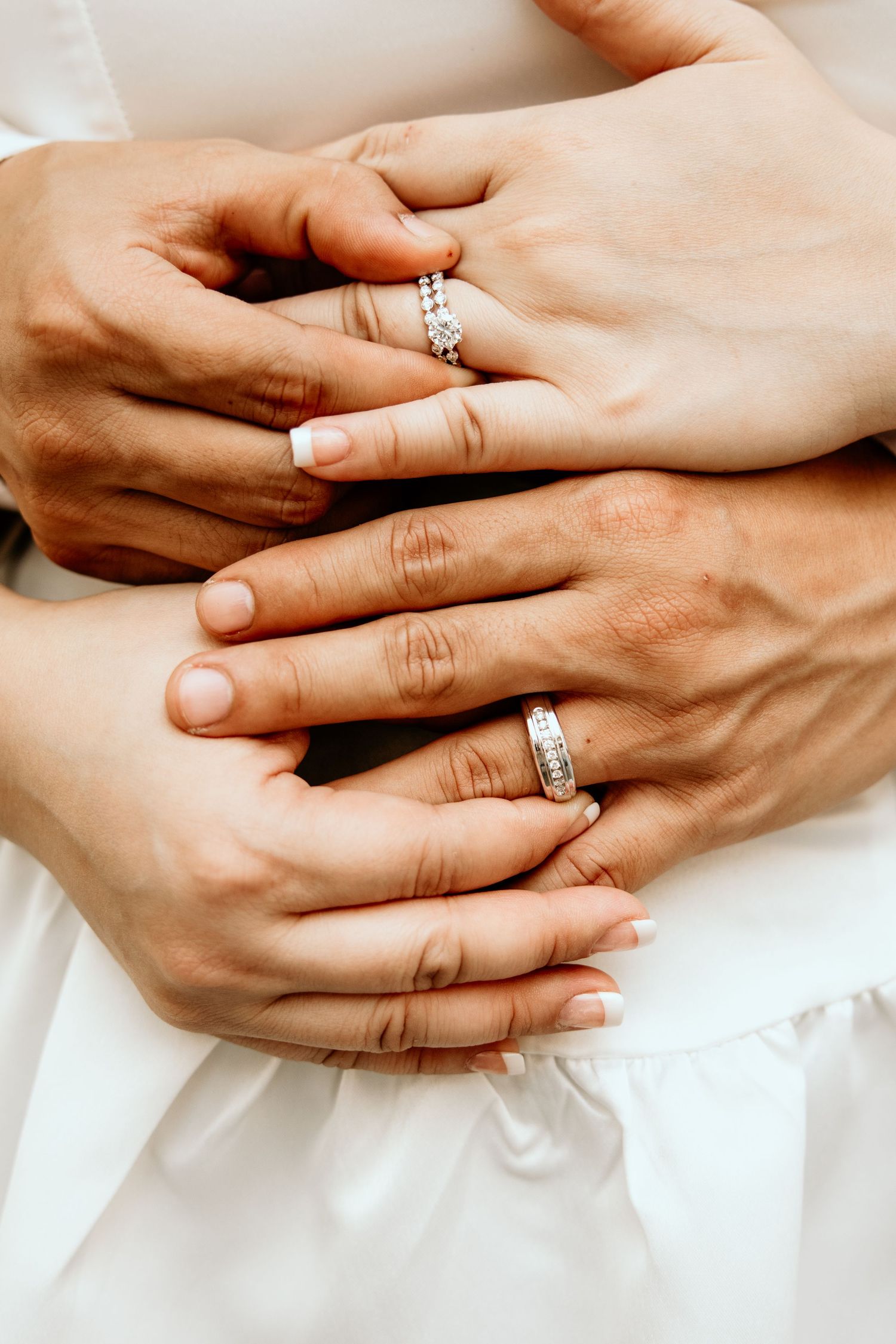 Close-up of bride and groom's hands with matching diamond wedding bands resting on white bridal gown.