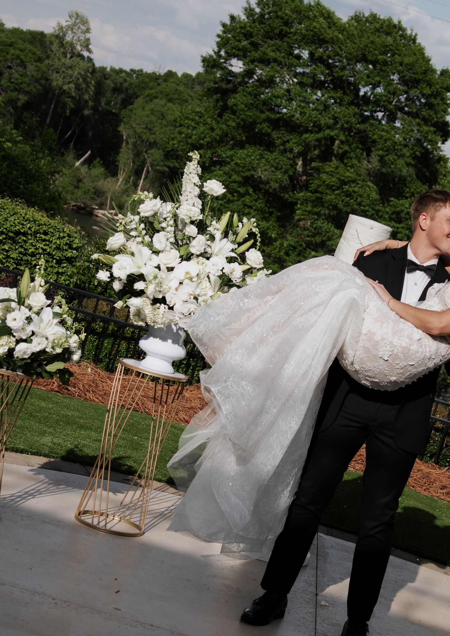 Groom carries bride in white lace gown past elegant white floral urns at outdoor wedding venue.