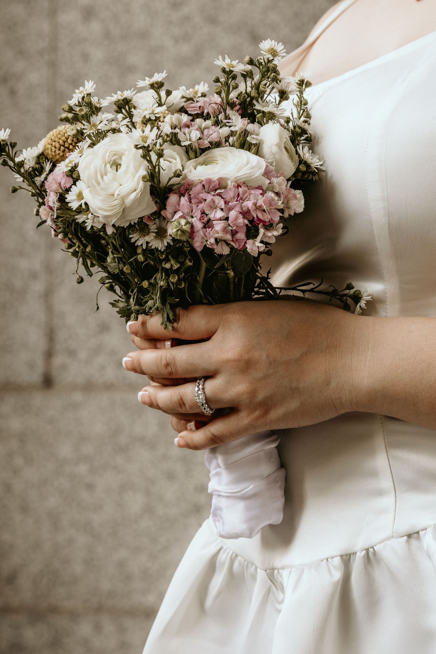 Bride holding a beautiful wildflower bouquet with white ranunculus and pink blooms, wearing a diamond ring and white dress.