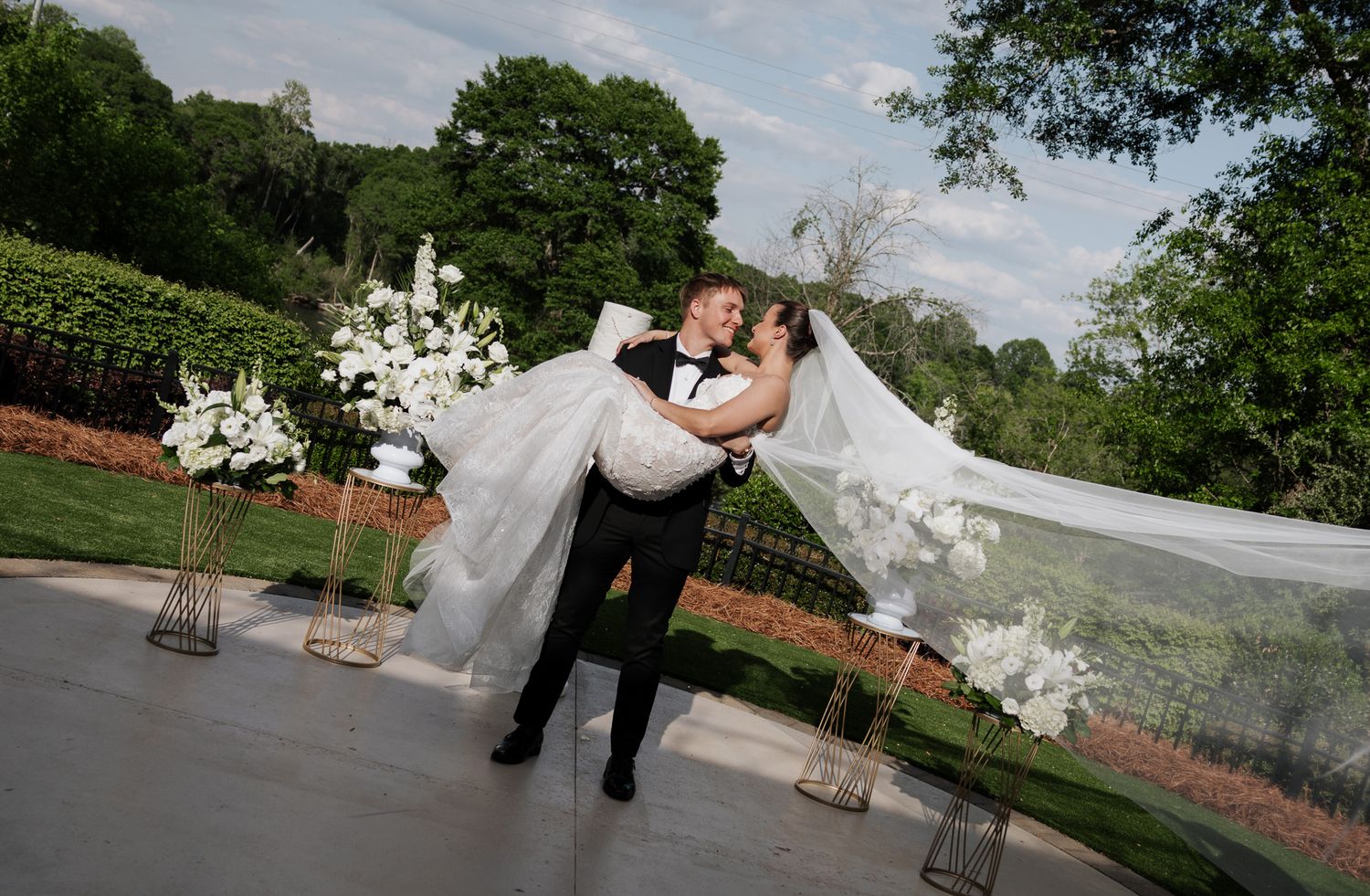 Groom carries bride in arms on outdoor terrace with white floral arrangements and scenic greenery backdrop.
