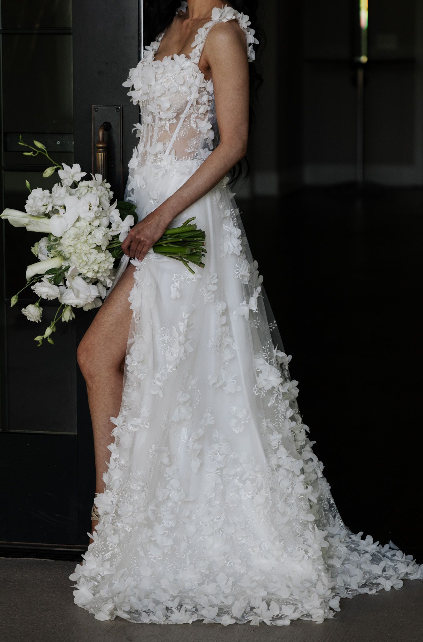 Bride in floral applique wedding gown with thigh slit holds white flower bouquet outdoors.