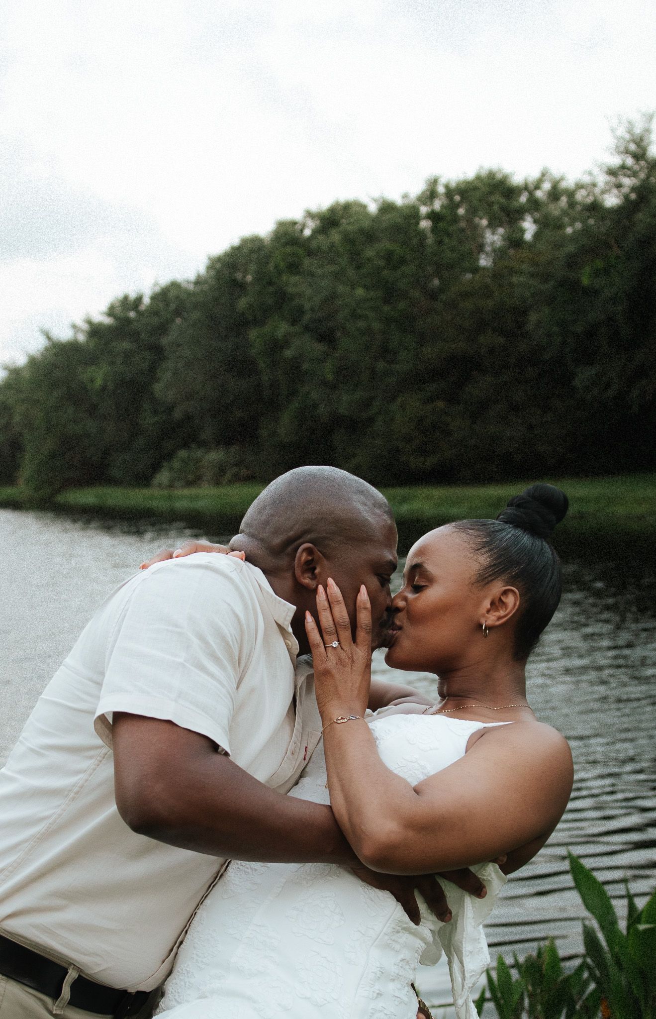 A joyful couple shares a romantic kiss outdoors near a serene lake surrounded by lush green trees.