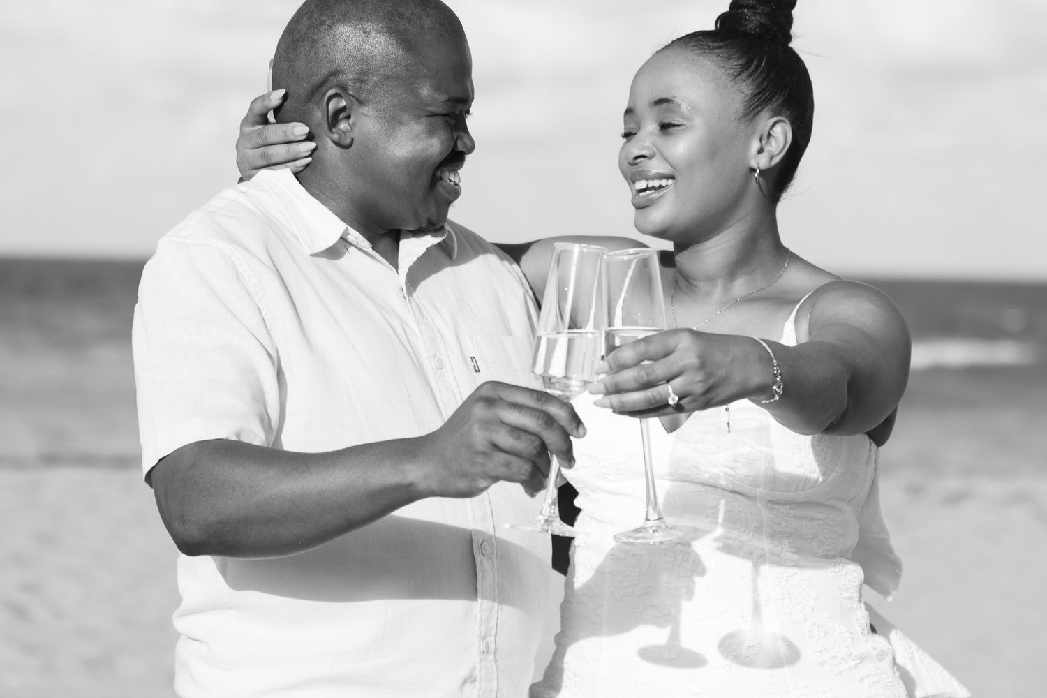 Black and white photo of a couple toasting with champagne glasses on a beach, smiling at each other.
