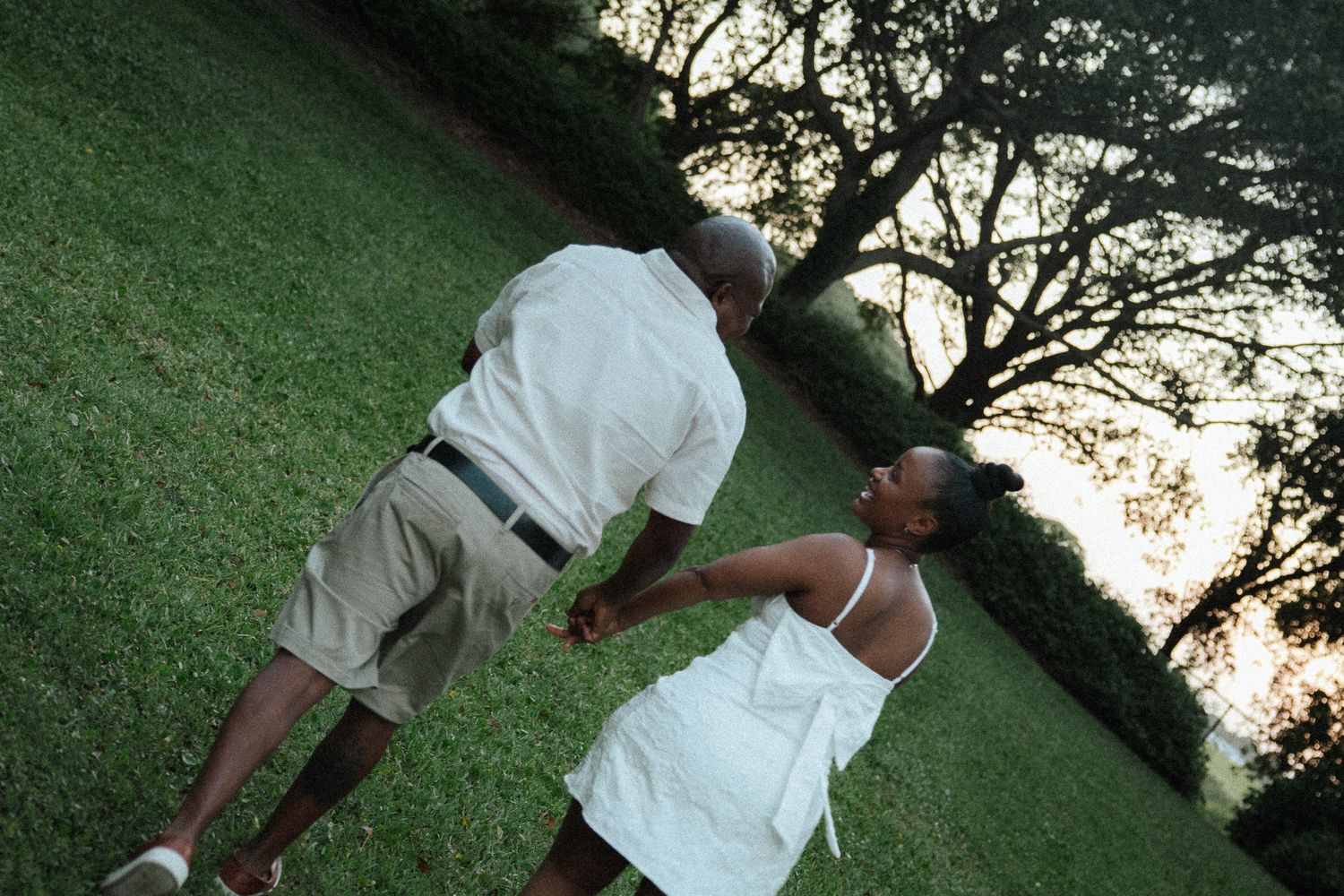 A happy couple walks hand in hand across a lush green lawn at sunset near a large tree.