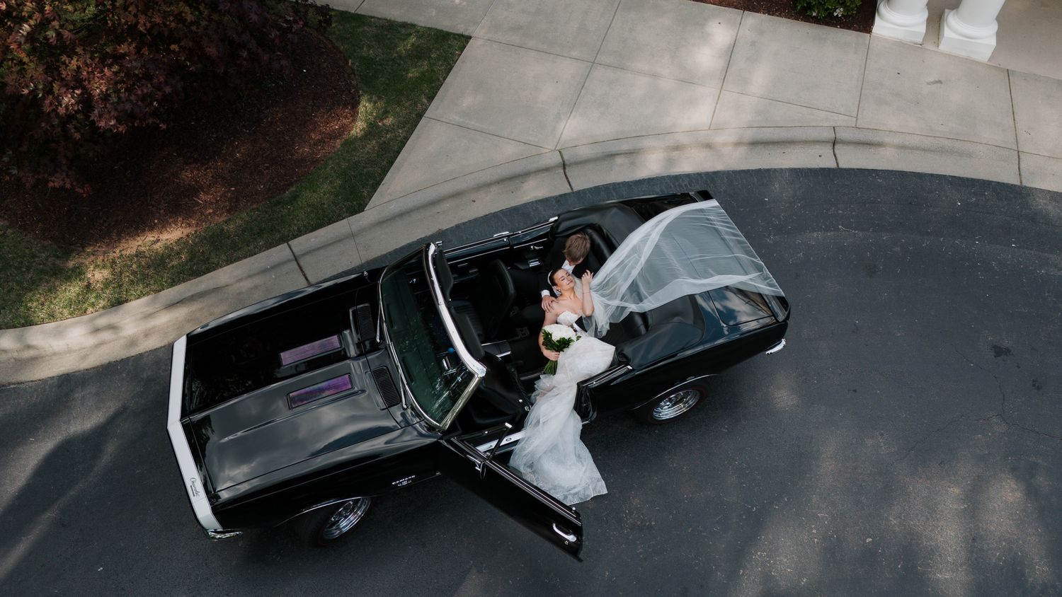 Aerial view of bride and groom embracing beside a classic black convertible on a driveway.