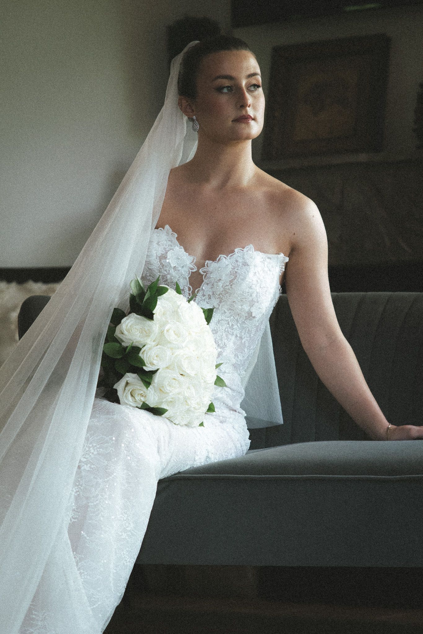 Graceful bride in a strapless lace wedding dress with long veil holds white rose bouquet while seated on a sofa.