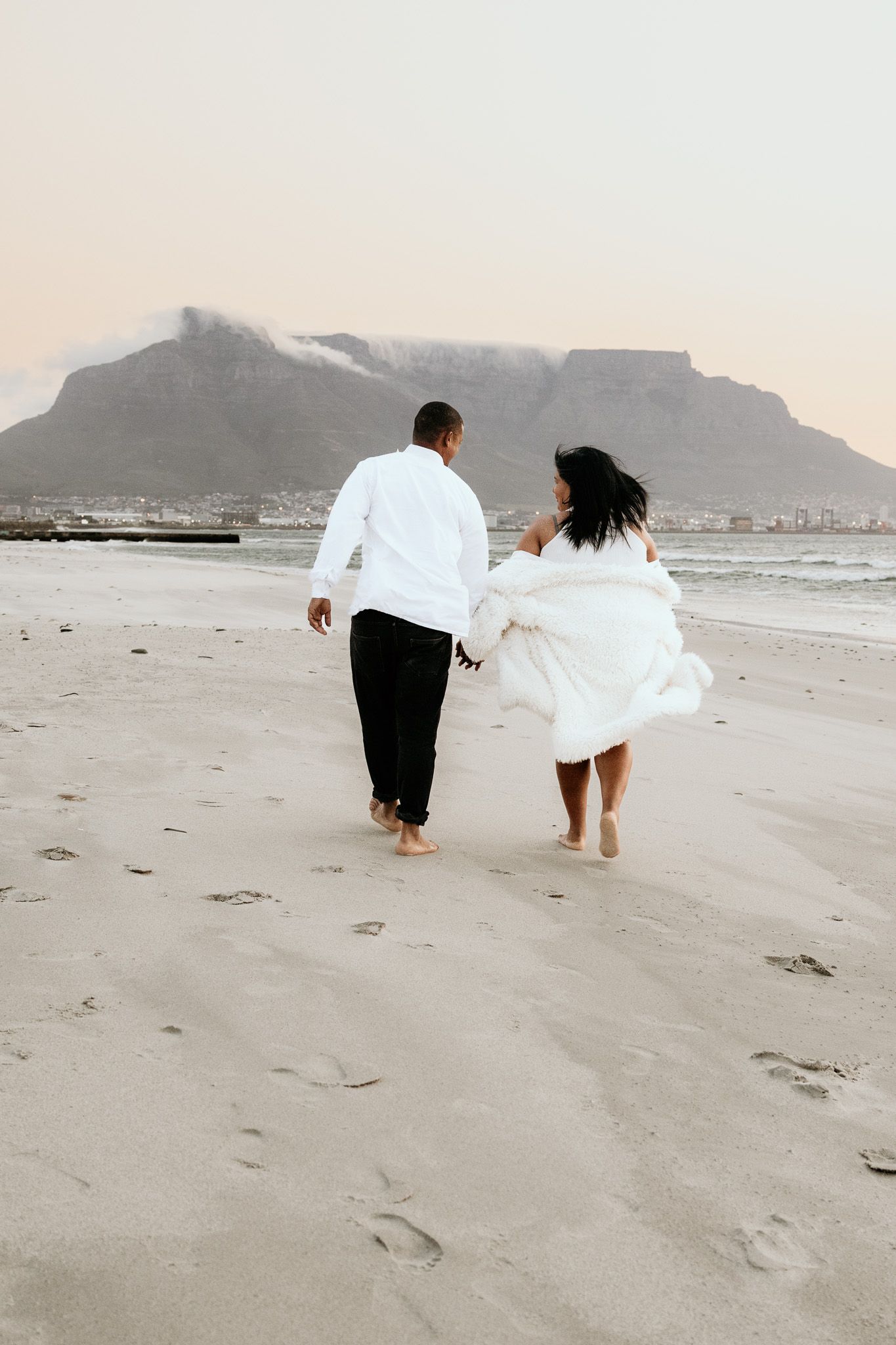 Couple walking barefoot on beach toward Table Mountain, bride in flowing white dress at sunset.
