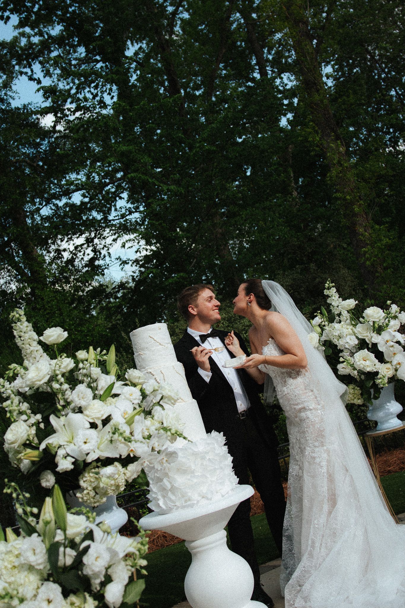 Bride playfully feeding groom beside a stunning white tiered wedding cake surrounded by white floral displays outdoors.