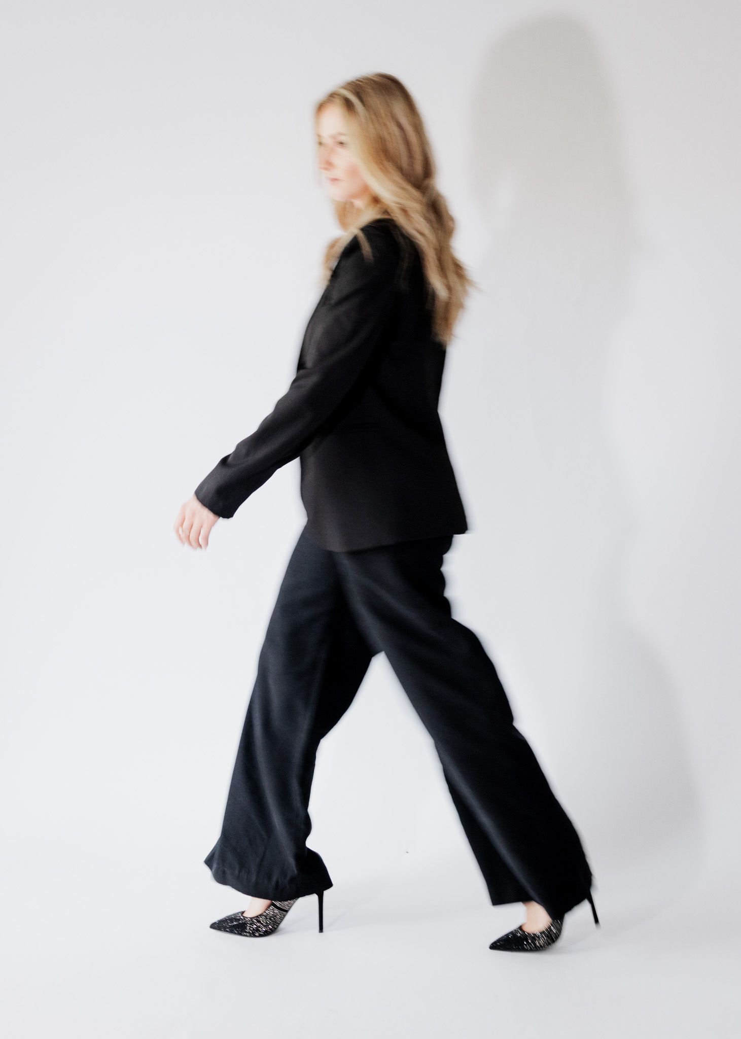 Blonde woman walking in black blazer and wide-leg trousers with stiletto heels against white background.