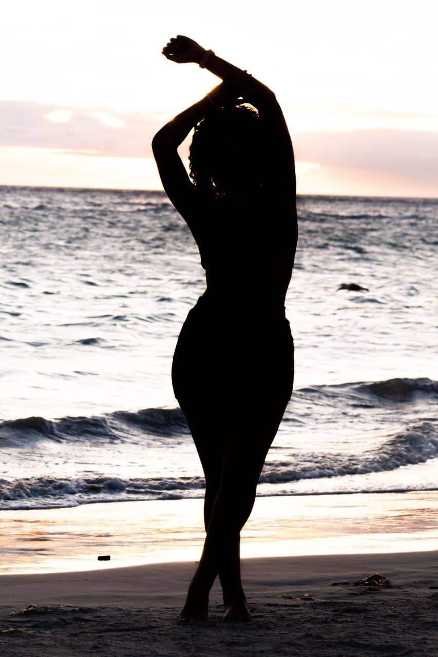 Silhouette of a woman posing gracefully on a beach at sunset with ocean waves in the background.