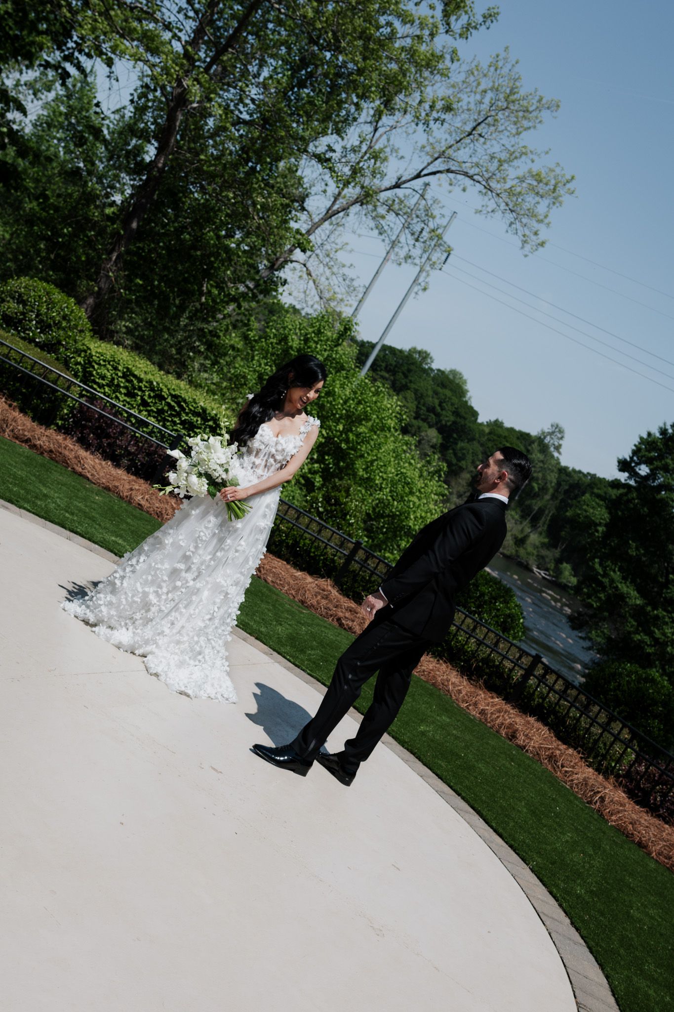 Bride in lace gown holds white bouquet facing groom in black tux on curved garden path.