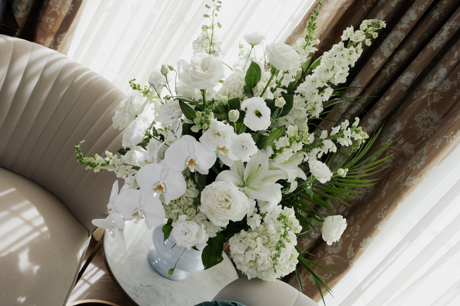 Lush white wedding bouquet featuring orchids, roses, and greenery in a marble vase.