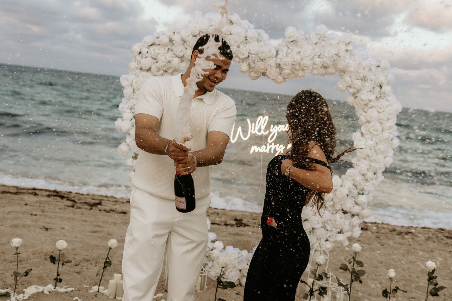 Beach marriage proposal with champagne celebration in front of a white rose heart arch.