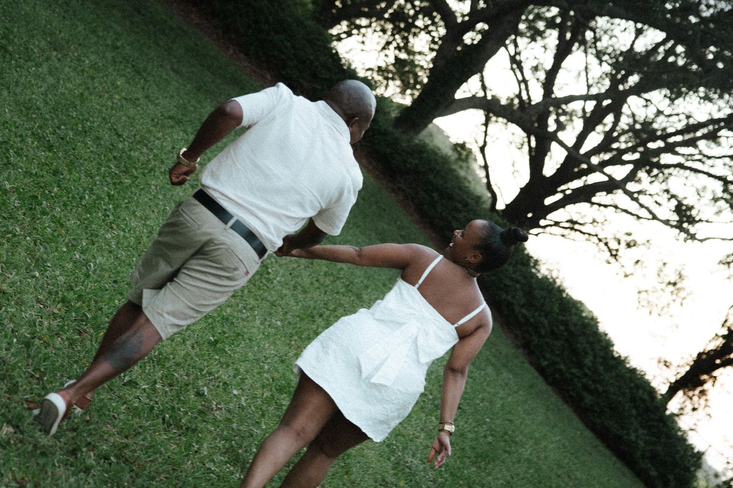 An aerial view of a couple holding hands while walking across a vibrant green grassy park at dusk.