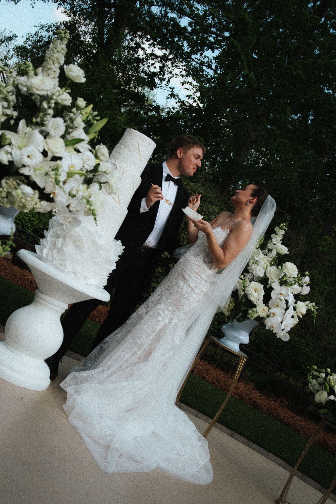 Bride and groom share wedding cake outdoors surrounded by white floral arrangements and lush greenery.