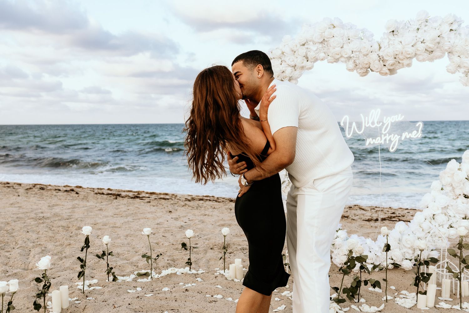 Couple sharing a kiss under a white balloon heart arch on a beach surrounded by white roses.