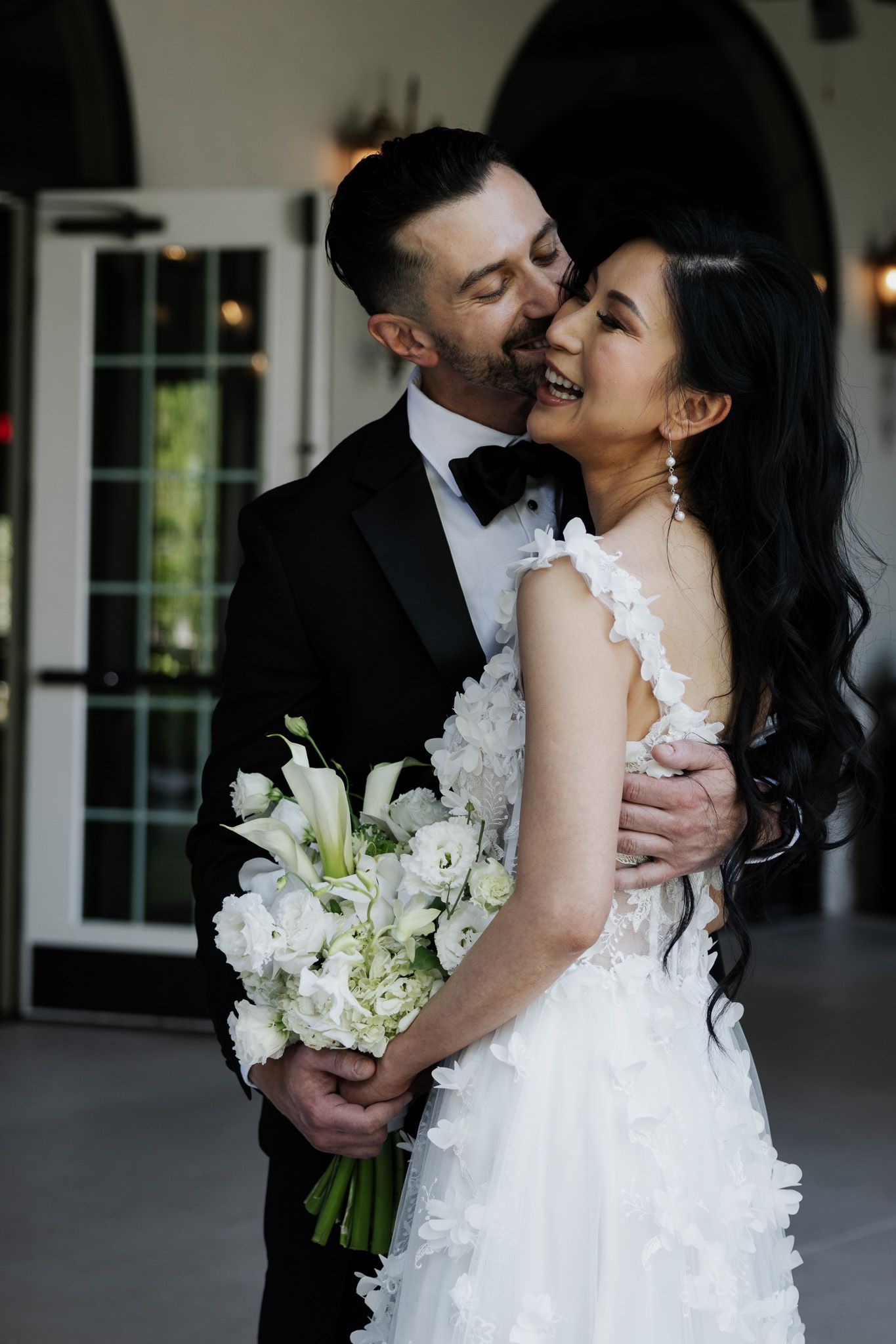 A bride in a white floral gown holds a bouquet while a groom in a black tuxedo embraces and kisses her cheek.