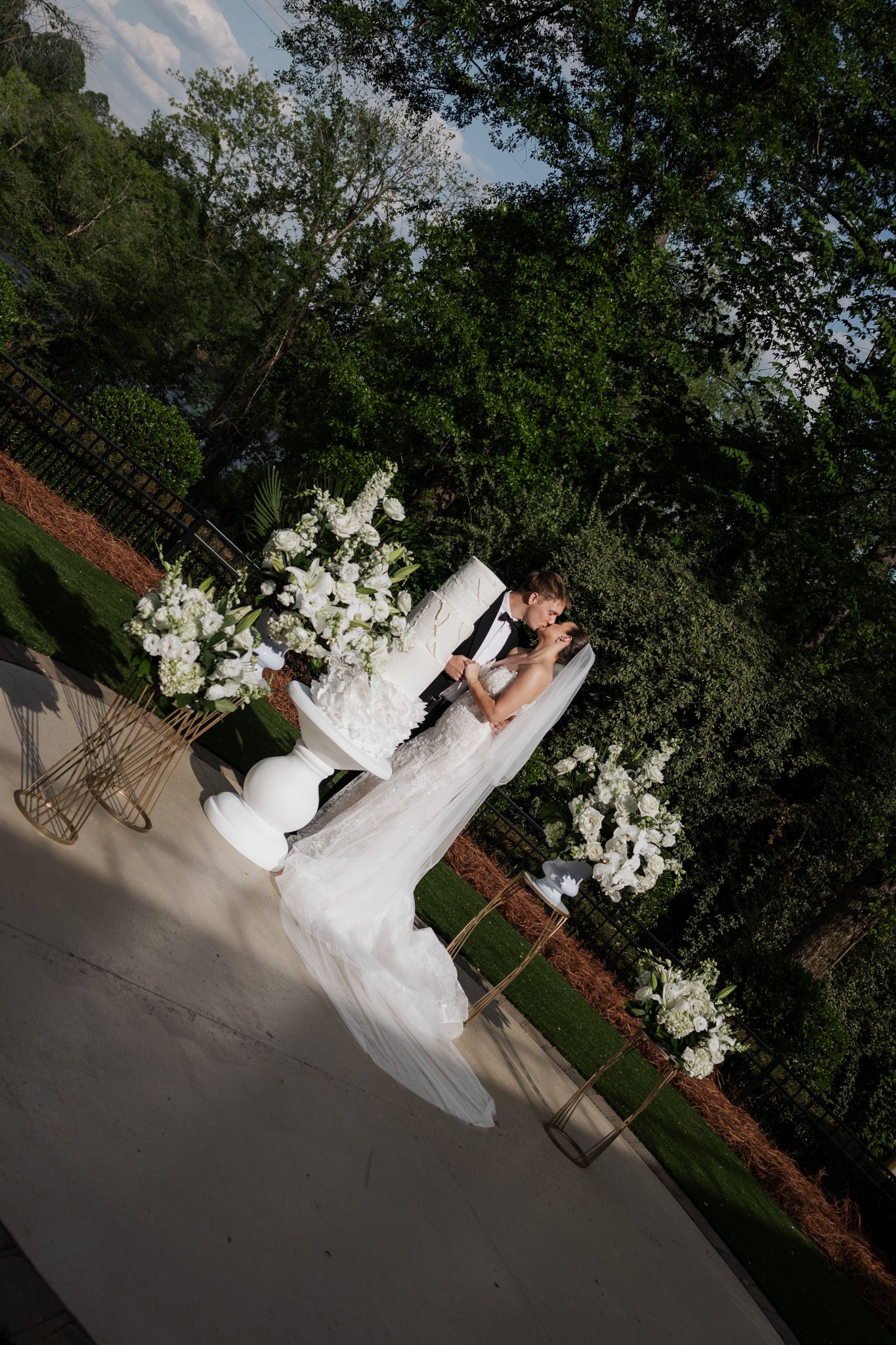 Aerial view of bride and groom kissing beside tall white floral arrangements in lush garden setting.